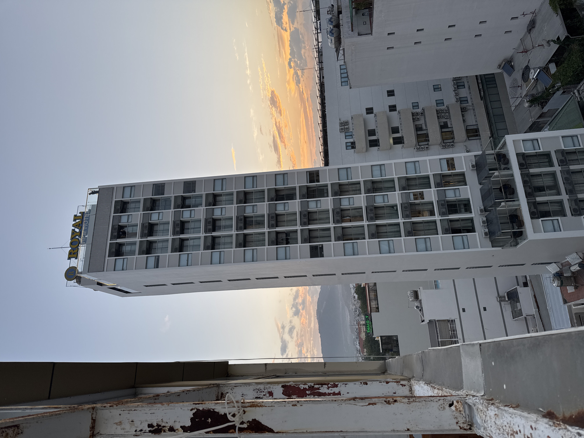A tilted, medium-shot photograph captures an urban landscape in Nha Trang, Vietnam, during what appears to be late afternoon or early evening, evidenced by the warm, orange-tinted sky and scattered clouds. In the foreground, to the left, a weathered, rust-colored metal railing with peeling white paint creates a distressed barrier. Dominating the center is a tall, slender, multi-story white building with a prominent yellow "ROYAL" sign at its apex, likely a hotel, identified by the "NEW HOTEL" text below. The building's facade is characterized by rows of windows, many with dark curtains drawn, and air conditioning units mounted on the exterior.
To the right of the central building, several other white buildings of varying heights are visible. One building has multiple balconies and numerous air conditioning units. Another, further to the right, features a simpler facade with regularly spaced small windows. A wider view reveals a hazy mountain range in the distance. The rooftop of the "ROYAL NEW HOTEL" has a railing and a solitary silhouette of a person standing near the edge, looking out. The overall impression is one of a dense urban environment with a striking architectural centerpiece against a backdrop of a colorful sky and distant natural scenery.