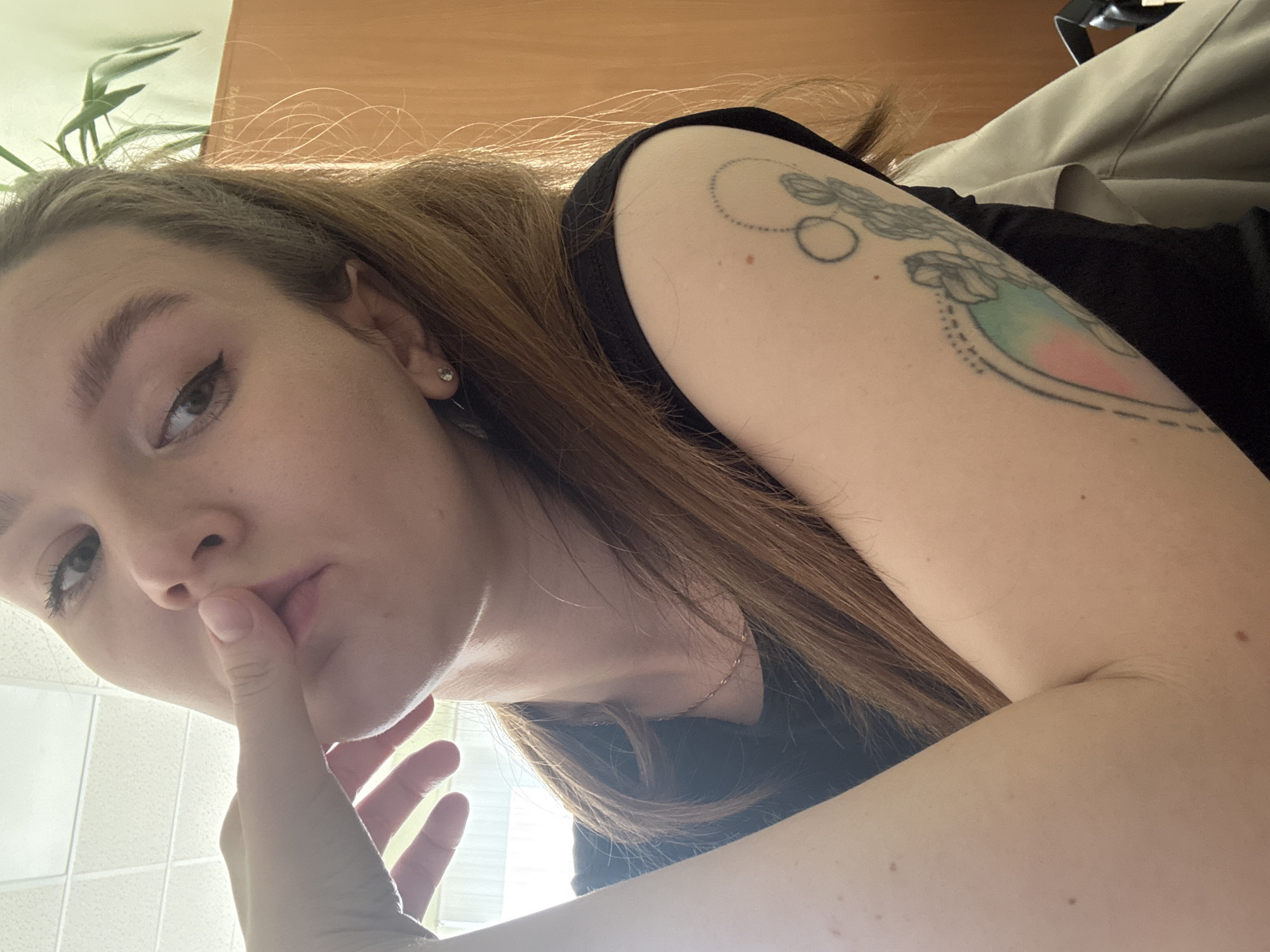 A young Caucasian woman with long, light brown hair is shown from the chest up. She is wearing a black top and a delicate necklace. Her right hand is touching her lips, and her left arm is resting on a light-colored jacket. She has a tattoo on her left arm. The background shows a ceiling with fluorescent lighting, a light-colored wall, and a wooden cabinet. The overall lighting is bright, possibly from natural light and overhead fixtures. The image appears to be taken indoors, likely in an office or study, in Izhevsk, Russia.