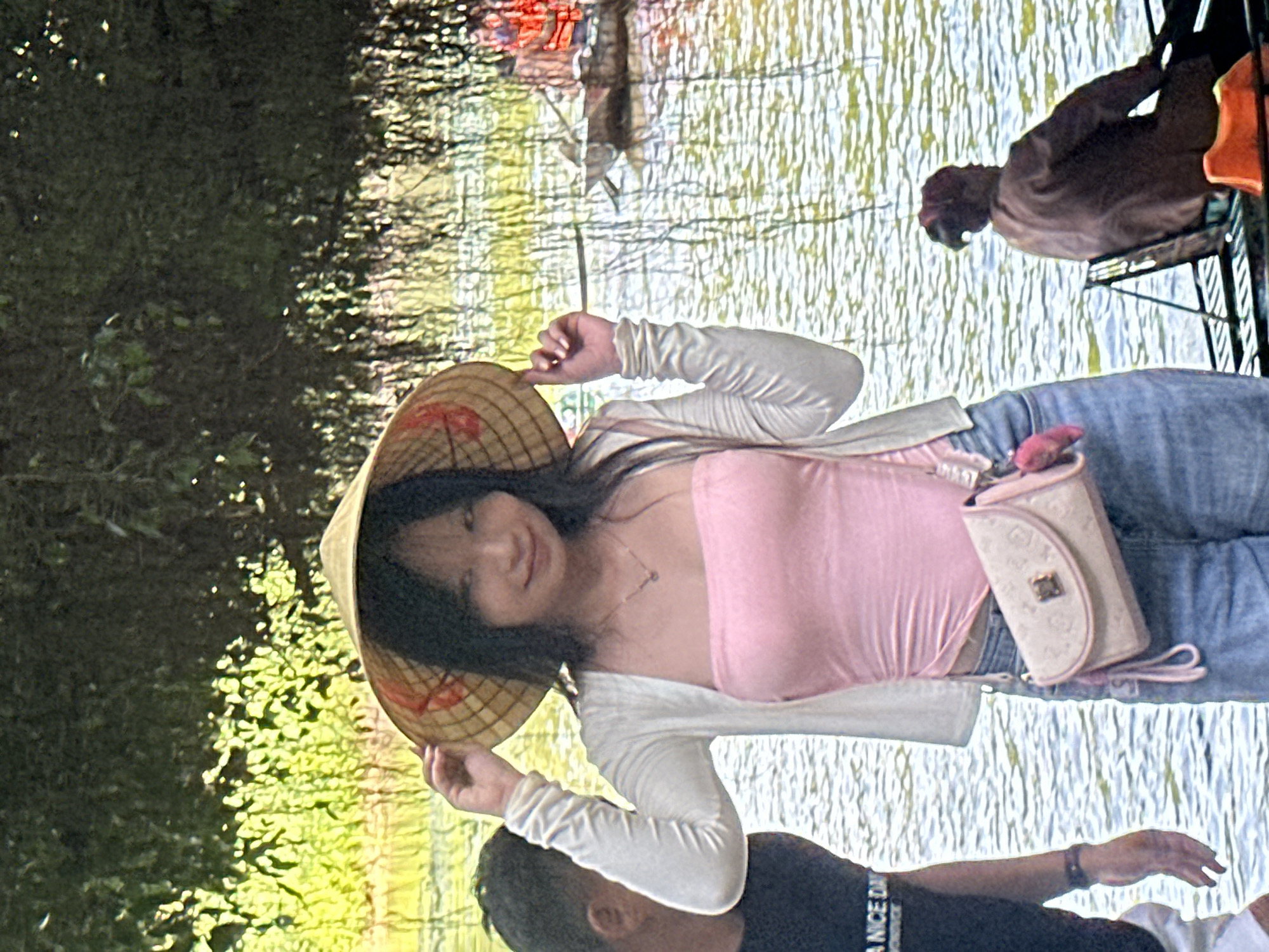 In Yên Sơn Ward, Vietnam, a young woman stands by a body of water, wearing a conical hat, a pink strapless top, a white cardigan, and jeans. She has a small pink purse hanging from her shoulder. To her left, partially visible, is a person wearing a black shirt. In the background, a few boats are on the water, with one occupied by a person wearing an orange life vest and another by a person rowing. The environment appears to be a natural setting with trees and foliage surrounding the water.