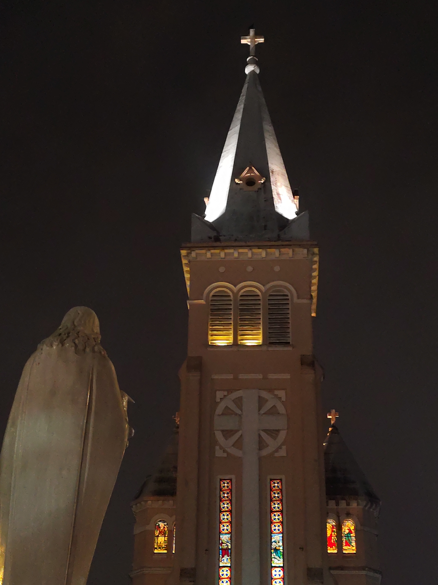 The image presents a dramatic nighttime view of a church tower and a statue, likely in Da Lat, Vietnam. In the left foreground, a large, light-colored statue is seen from the back, draped in robes, suggesting a religious figure like Mary.
Dominating the right and central background is a tall church tower, characterized by its warm, light brown or peach coloration, reminiscent of the Da Lat Cathedral. The tower culminates in a sharp, white spire, which is brightly illuminated by upward-pointing lights. A cross crowns the very top of the spire. Below the spire, the tower features a section with three arched windows, fitted with louvers, from which a warm yellow light emanates. Further down, a large circular emblem with a cross-like design is visible. The lower part of the tower displays several tall, narrow stained-glass windows, glowing with vibrant colors and depicting intricate patterns and what appear to be religious figures. A smaller dome or tower, also topped with a cross, is faintly visible behind the main structure.
The scene is clearly set at night, with the sky appearing uniformly dark and featureless. The artificial lighting dramatically highlights the architectural details of the church and the contours of the statue, creating a serene and majestic atmosphere. No discernible text is visible in the image.