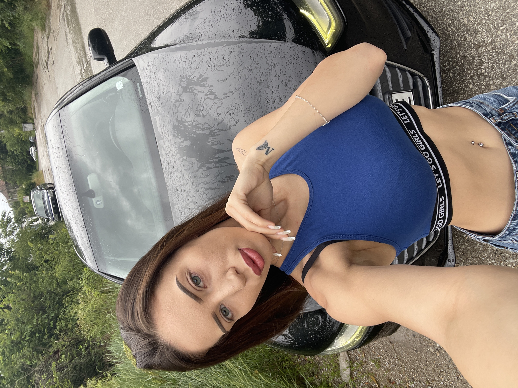 The image shows a young woman leaning against a black car. She is wearing a blue crop top and denim shorts. Her hair is dark brown and she has striking green eyes. She is wearing makeup, including lipstick and eyeliner. On her left wrist, she has a delicate silver bracelet and a small butterfly tattoo. She also has a belly button piercing.
The car appears to be wet, suggesting it might have recently rained or is raining. The background shows greenery, possibly trees and grass, and a paved surface that could be a road or parking lot. The lighting suggests it is daytime. The woman's pose is relaxed, with one hand touching her face, and her expression is direct and confident. The text "LET'S GO GIRLS" is visible on the elastic band of her crop top. The overall scene feels casual and outdoorsy.