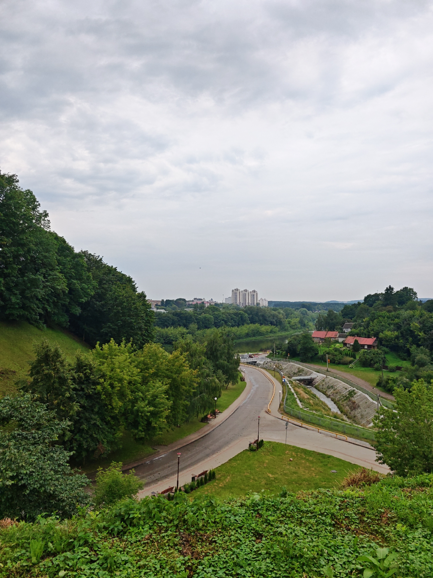The image depicts a panoramic view of a landscape with a city in the distance, located in Hrodna, Belarus. In the foreground, lush green vegetation covers a grassy slope. A paved road curves through the middle ground, flanked by green grass and trees. A concrete embankment with a channel for water runs alongside the road.
Further in the distance, a river is visible, bordered by trees and several small buildings with red roofs. Beyond the river, a cluster of tall, modern apartment buildings stands against the backdrop of a cloudy sky. The overall scene is serene and green, showcasing a blend of natural landscape and urban development. There are no people visible in the image. The lighting suggests an overcast day.