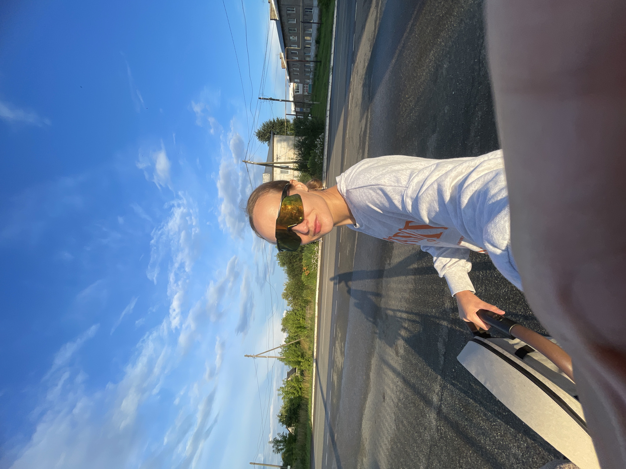 In Подшивалово, Russia, a young woman with blonde hair pulled back and wearing large, reflective sunglasses, a grey sweatshirt with orange lettering, stands on a paved road. She is holding onto the handle of a light-colored stroller. The sun is low in the sky, casting long shadows across the road. In the background, there are buildings, trees, and utility poles under a blue sky with scattered clouds. The image appears to be a selfie, taken by the woman herself.