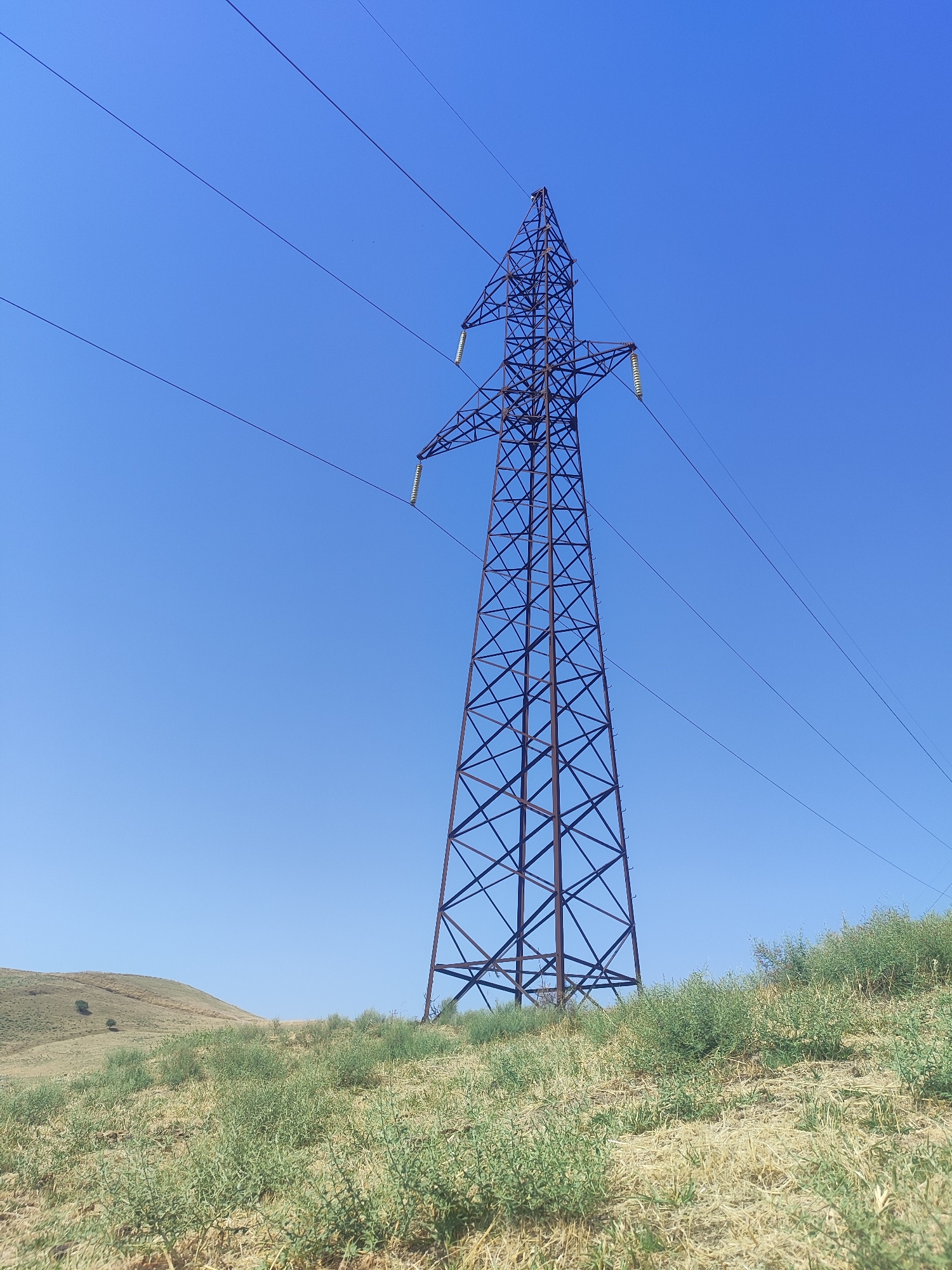 This is a full shot image of a power line tower in a rural landscape under a clear blue sky. The power tower is made of dark metal lattice and stands tall against the sky, with several wires extending from it. The ground in the foreground and midground is covered in dry grass and low green shrubs, with a gentle, sloping hill rising to the left. The image appears to be taken on a sunny day. The location is identified as Sultanabad, Tajikistan.