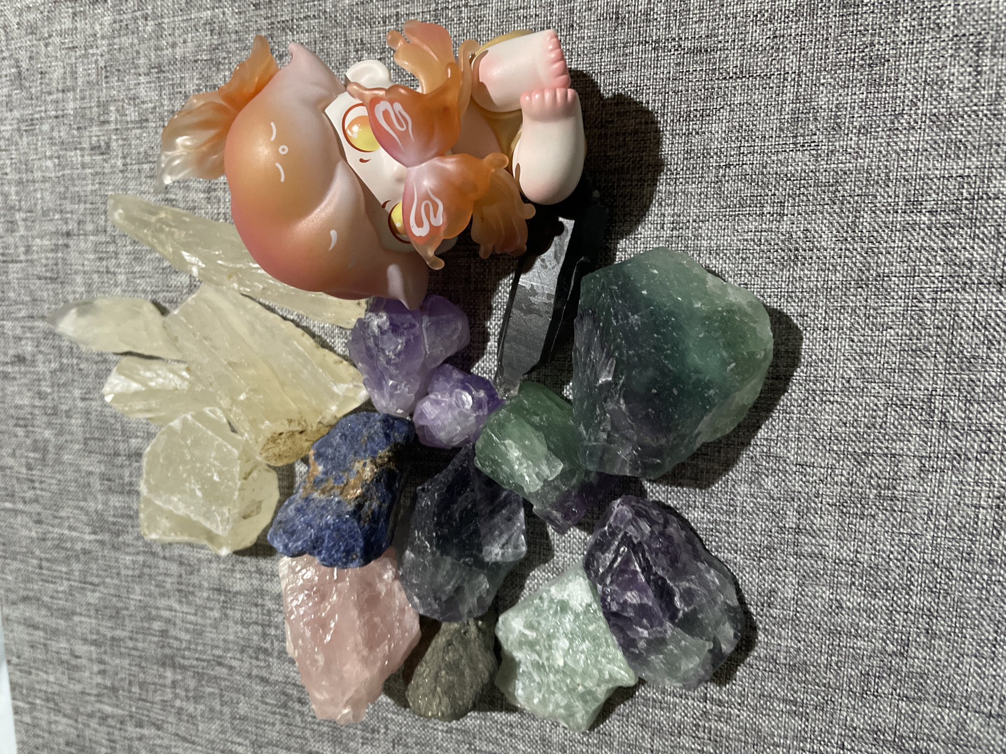 A collection of natural mineral specimens and a stylized collectible figurine are arranged on a textured, light gray woven fabric surface in Davao City, Philippines. The minerals, roughly cut and unpolished, include several elongated, translucent pale yellow formations resembling selenite or calcite. Other specimens feature a rounded pink stone (possibly rose quartz), a dark blue stone with brown flecks (likely sodalite), various irregular chunks of purple and green minerals (consistent with fluorite or amethyst), and one dark gray, possibly metallic specimen. On the right side of the arrangement, a peach-toned figurine with a smooth, rounded head and stylized closed eyes and mouth holds a smaller, winged figure. The larger figurine has translucent, peach-colored fin-like extensions from its head, while the smaller figure displays large yellow eyes on its body and translucent orange butterfly-like wings. Shadows are cast predominantly to the left and bottom of the objects, indicating an overhead light source.