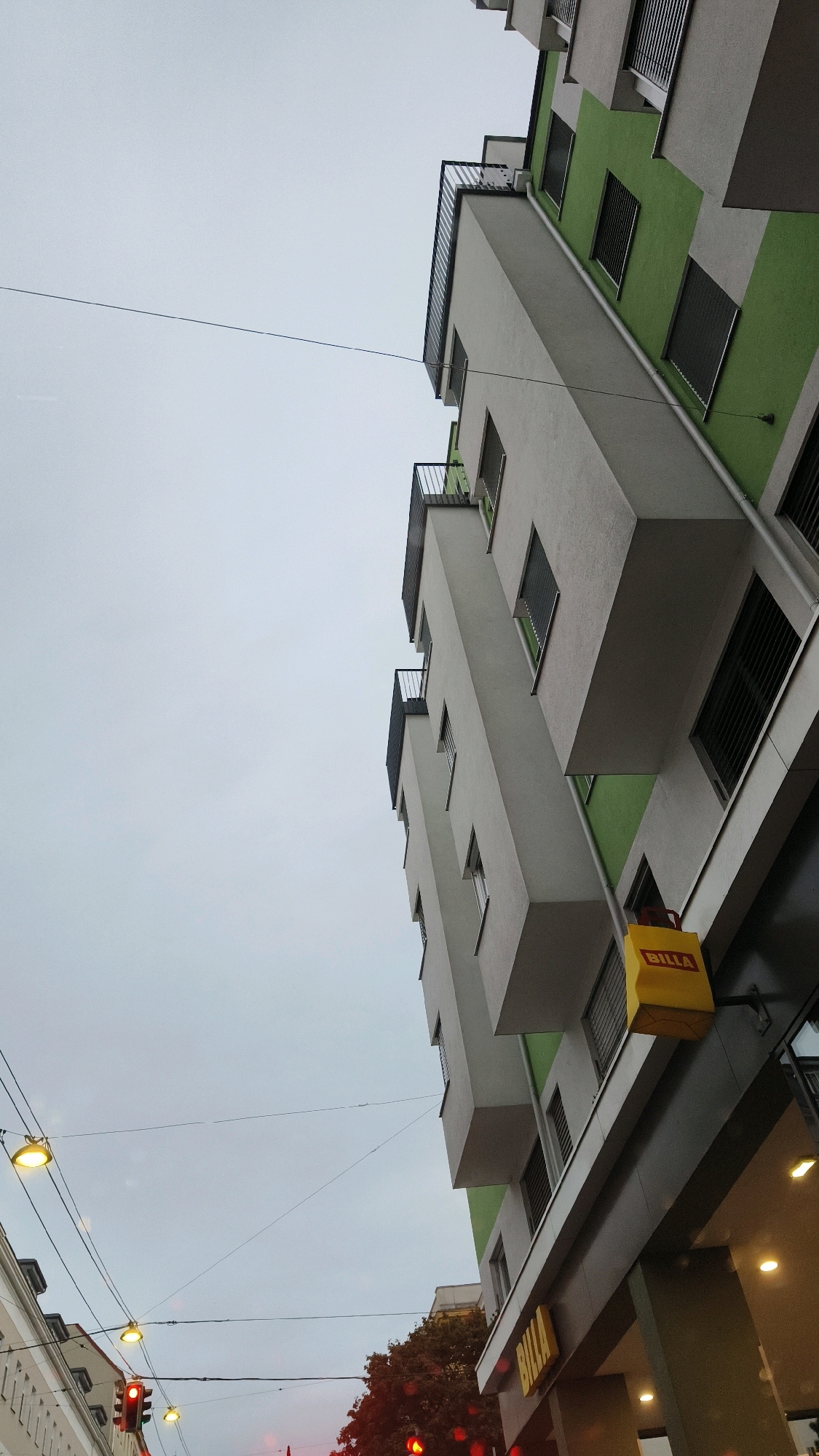 A low-angle, full-shot image captures a modern apartment building with green and white facades in Vienna, Austria. The building features multiple balconies with dark railings and large windows with dark shutters. A yellow sign with the red "BILLA" logo is mounted on the side of the building, indicating a supermarket. Below the building, the entrance to the BILLA store is visible with lit interior spaces. Overhead, electrical wires crisscross the frame, with several street lamps and a traffic light visible in the lower left corner, showing a red signal. The sky is overcast and grey. The image is taken from street level, looking upwards towards the building.