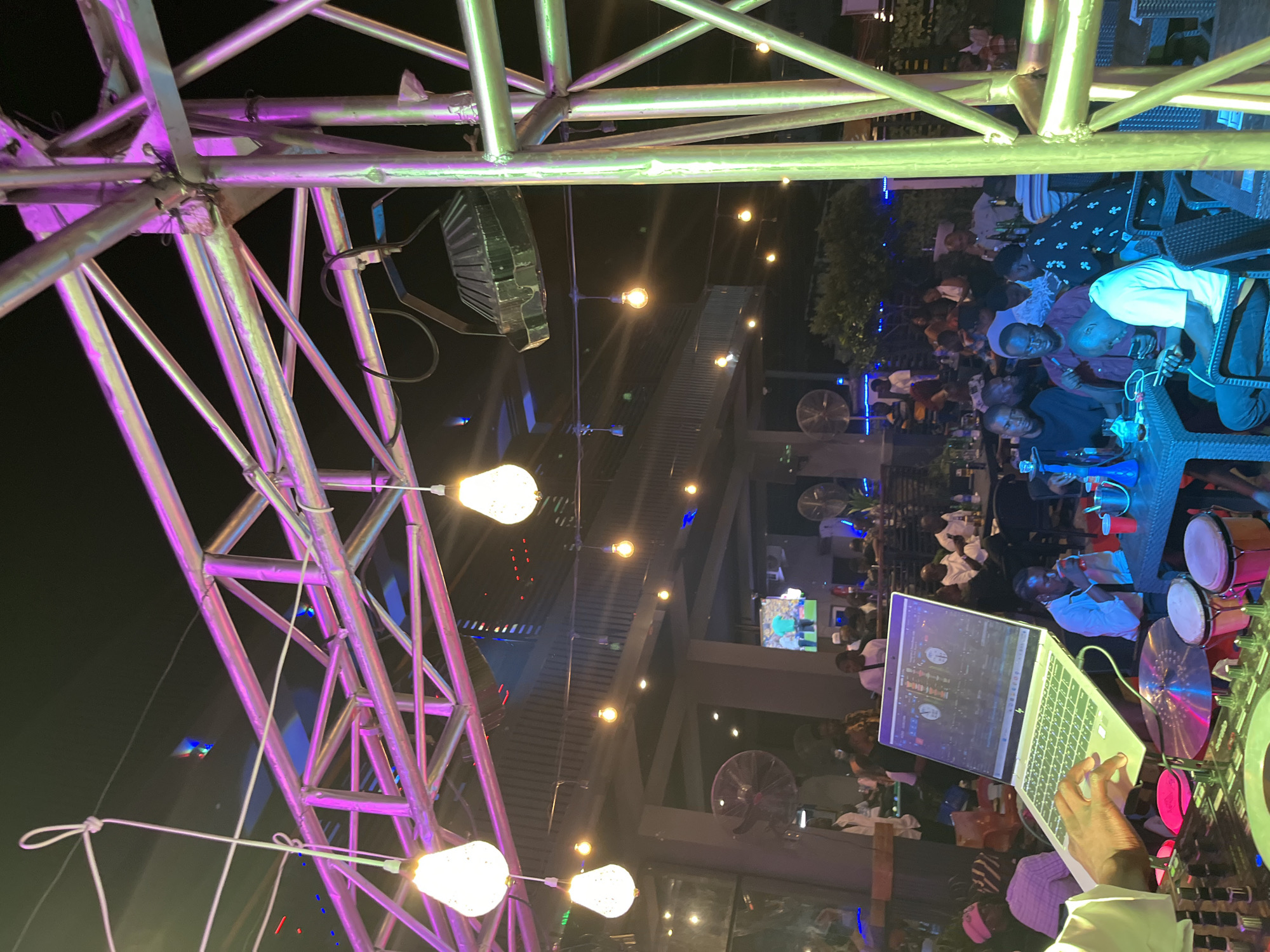 The image shows an outdoor nighttime scene at a venue in an unknown city. The foreground is dominated by a DJ setup, including a laptop, DJ controller, and hand-held drums. A person's hand is visible, operating the laptop. In the midground and background, a crowd of people are seated at tables, some with drinks and hookahs. The venue appears to be lit by decorative lights, including pendant lamps and string lights, and is illuminated by purple and green stage lighting mounted on a metal truss structure. A large fan is visible on the left side of the image, and a television screen showing a sports broadcast is mounted on a wall in the background. The overall atmosphere suggests a lively social gathering or event.