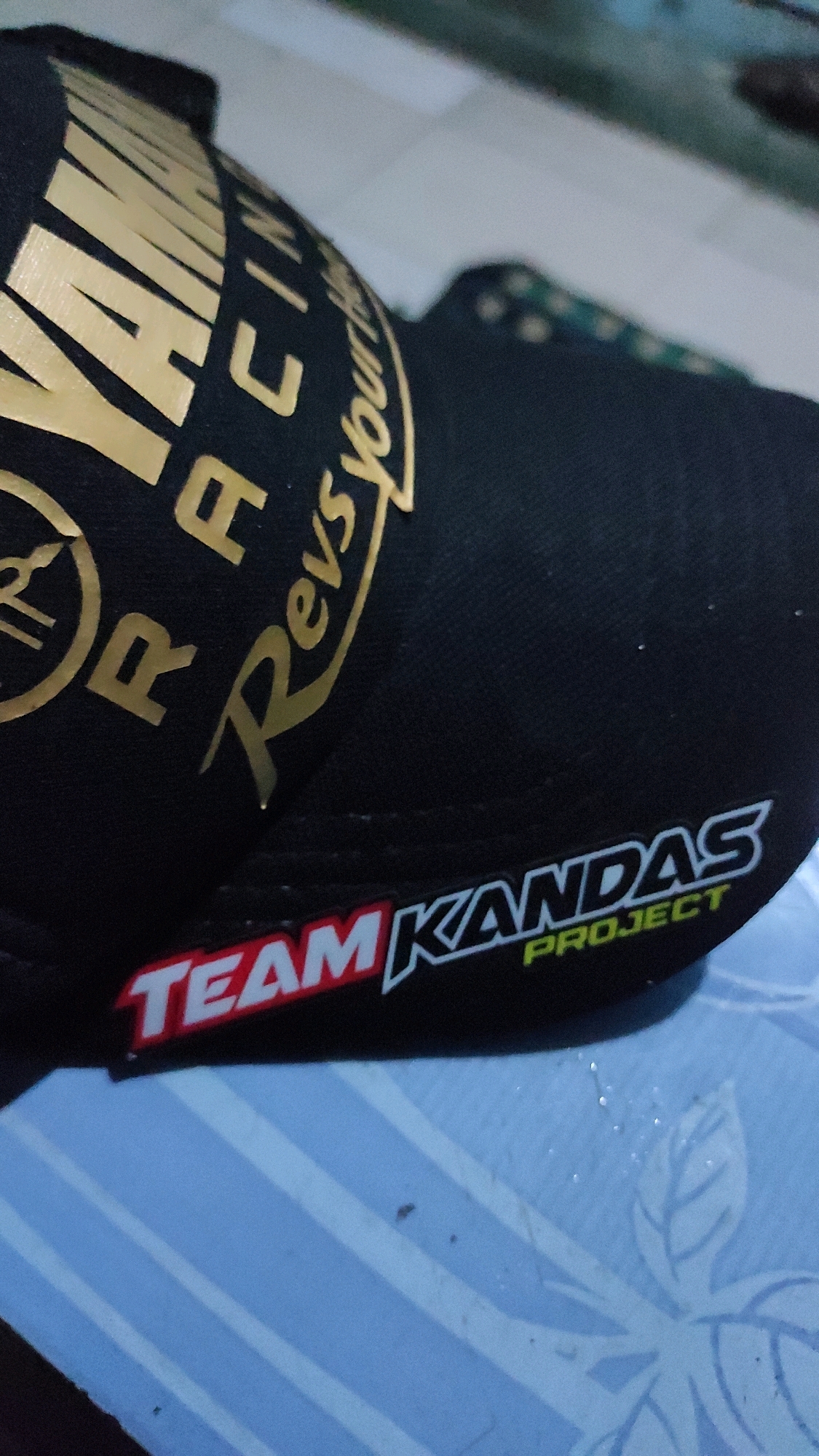 A black baseball cap with gold lettering is displayed. The cap has "YAMAHA RACING" in large gold letters and "Revs your heart" in a smaller, cursive gold font. A red and white patch on the brim reads "TEAM KANDAS PROJECT" in white lettering with a green outline. The cap is placed on a light blue surface with white stripes and a faint leaf pattern. The image is taken in Tanjungan, Indonesia.