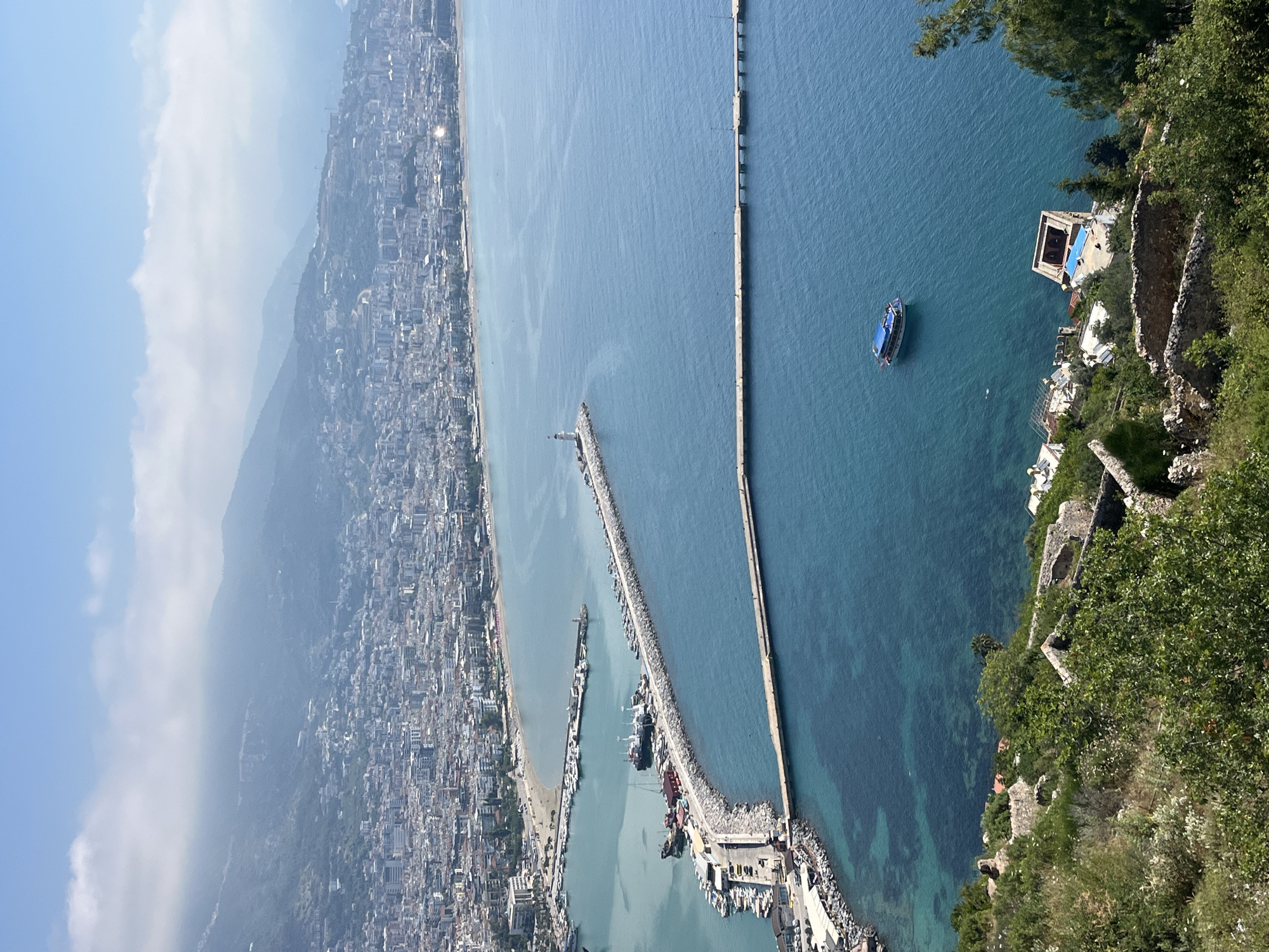 This is an expansive, high-angle view overlooking the coastal city of Alanya, Turkey, the Mediterranean Sea, and a bustling harbor, captured from a vantage point likely on a cliff or hill. The image is oriented vertically, showing the horizon from top-left to bottom-right if viewed as a landscape.
On the left side of the frame, a densely populated city stretches along the coastline, extending into the hazy distance where mountains rise. Numerous buildings and a sandy beach are visible, suggesting a vibrant urban and tourist area. The sky above the city and mountains is bright and clear with some light haze.
The center of the image is dominated by the clear blue waters of the Mediterranean Sea. A large, active harbor is prominently featured, protected by several long breakwaters. One main breakwater, lined with boats, extends into the sea, culminating in a small lighthouse. Numerous small to medium-sized boats are moored within the harbor, indicating a mix of fishing, commercial, and recreational maritime activity. Further out, a very long, straight breakwater runs parallel to the coast, creating an outer channel. A single small boat is seen navigating the open water beyond the main harbor.
In the immediate foreground, on the right side of the image, steep, rocky cliffs covered in lush green foliage descend towards the sea. Scattered amongst the greenery are remnants of old stone structures, possibly ruins, along with at least one modern building with a distinctive blue roof close to the water's edge. The water near the cliffs appears exceptionally clear, revealing the seabed.
The time of day is clearly daytime, with bright sunlight illuminating the entire scene, casting minimal shadows, suggesting late morning or early afternoon. The weather is sunny and clear, though a light atmospheric haze is present over the distant city and mountains. No people are individually discernible due to the distance, and no visible text is present.