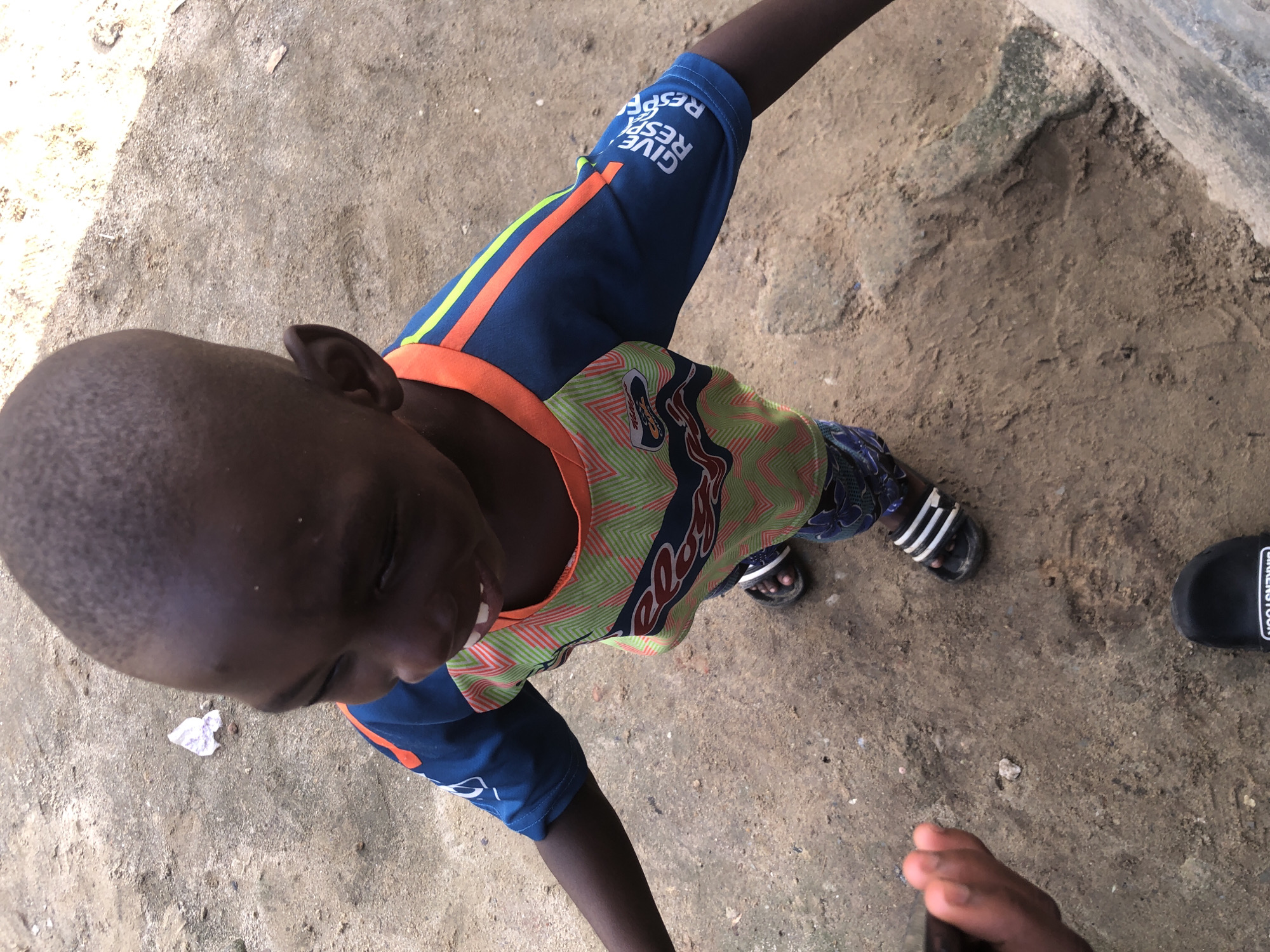 A young Black child, with a shaved head, is standing outdoors on a dirt ground in Ado, Nigeria. The child is wearing a short-sleeved t-shirt with a green and orange pattern, a blue t-shirt underneath, and patterned pants. The child is also wearing black sandals with white stripes. The child's right hand is visible, held up near their face. The ground is composed of dry soil and small stones. There is a piece of white and purple paper on the ground to the left of the child. A black sandal with "BIRKENSTOCK" visible on it is on the ground in the lower portion of the image.