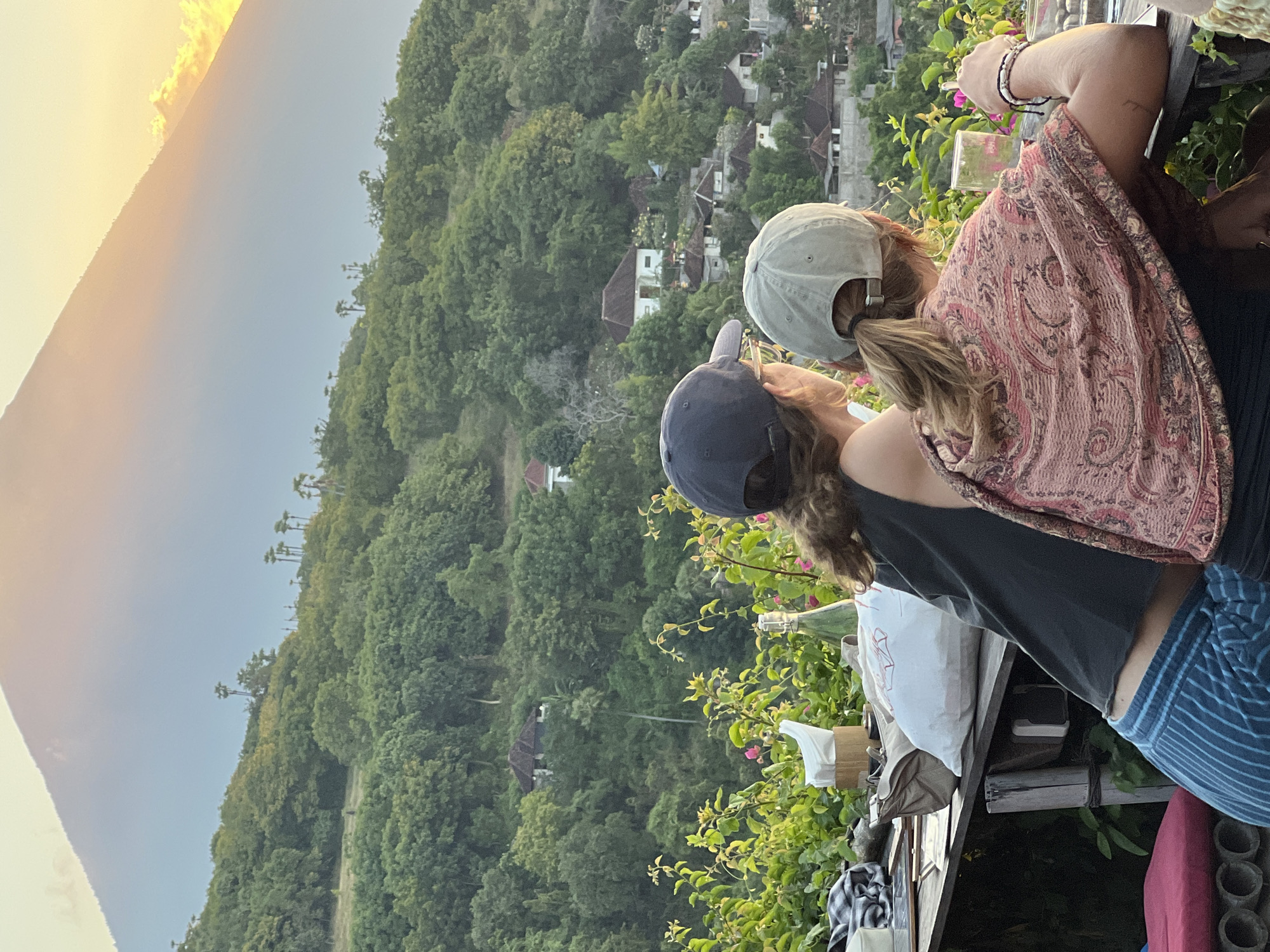 Two individuals, viewed from behind, are seated at a table overlooking a lush, green hillside dotted with homes. A large, imposing mountain dominates the background, partially obscured by a hazy, golden light, indicative of either sunrise or sunset. The person on the left wears a dark top and blue striped pants, with a dark baseball cap. The person on the right is adorned with a patterned shawl and a light-colored baseball cap. The table in front of them holds a napkin dispenser, a bottle, and other assorted items. The scene appears to be a scenic overlook in Abang, Indonesia, offering a tranquil view of the natural landscape and village life.