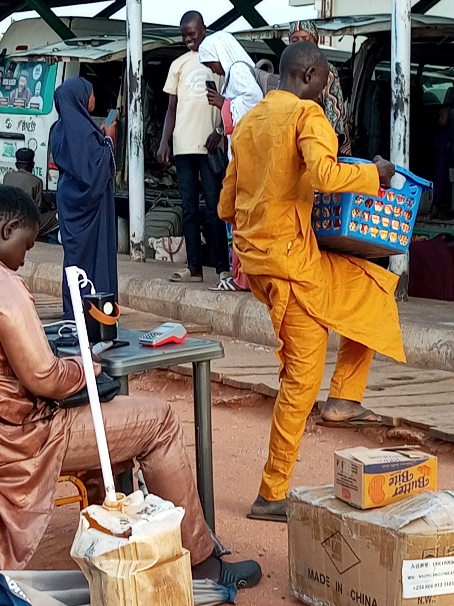 The image depicts an outdoor scene in the city of Gombe, Nigeria, possibly at a bus station or marketplace. In the foreground, a person dressed in a light brown outfit is seated, with a white pole and a portable charging unit on a small table in front of them. To the right, a man in a vibrant yellow traditional outfit is walking and carrying a blue plastic basket filled with items. Behind him, other individuals and luggage are visible, suggesting a transportation hub. The background features a white van with graphics on its side, and the overall environment appears to be dusty or unpaved.