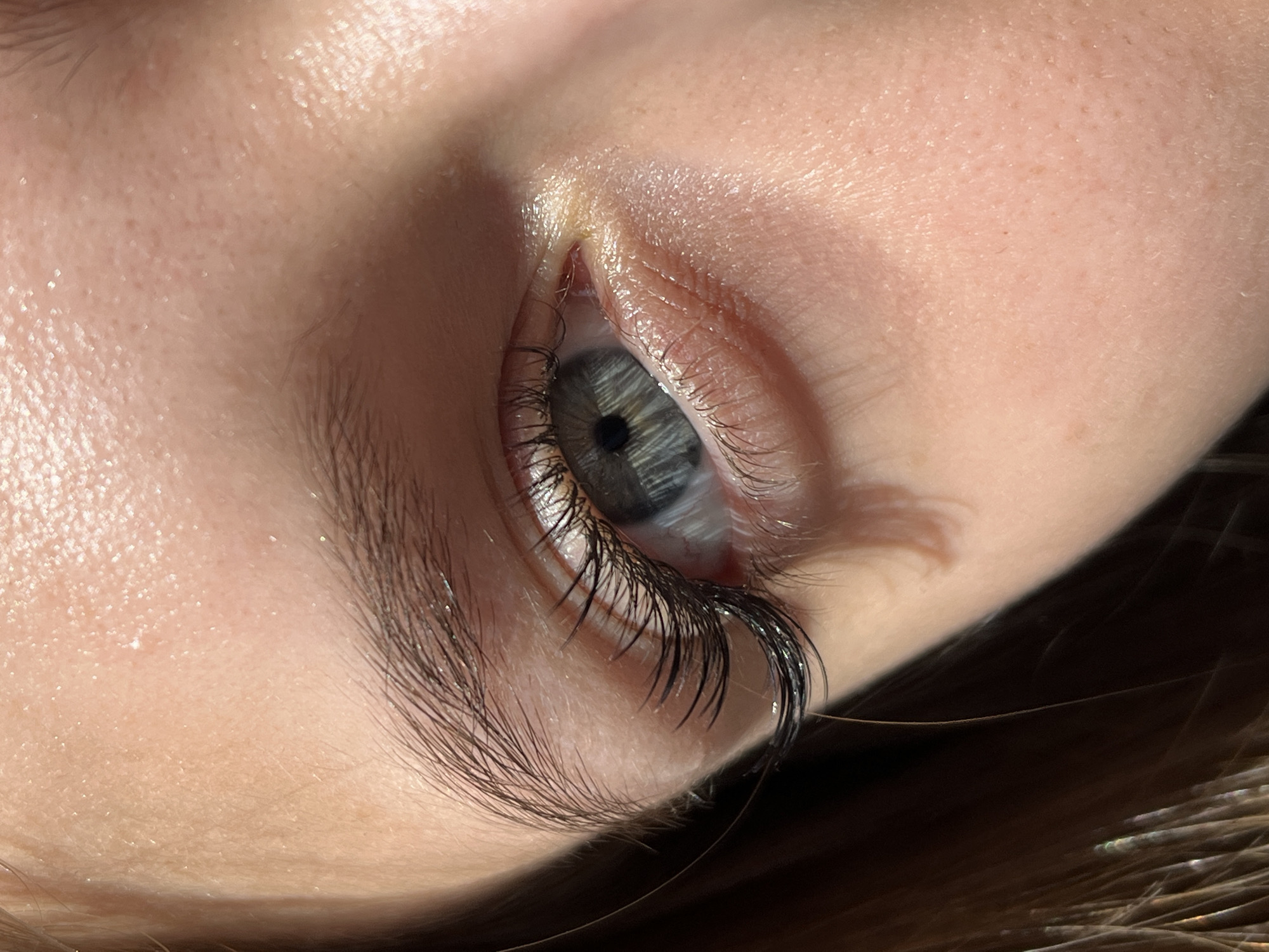 A close-up shot focuses on a human eye, presumably female, with long, dark eyelashes. The eye itself is a light blue-grey color. The surrounding skin appears fair, with a slightly oily sheen. The lighting is bright and casts shadows on the side of the face. Some strands of brown hair are visible near the bottom of the frame. There is no information to suggest that this image was taken in Фоки, Russia.