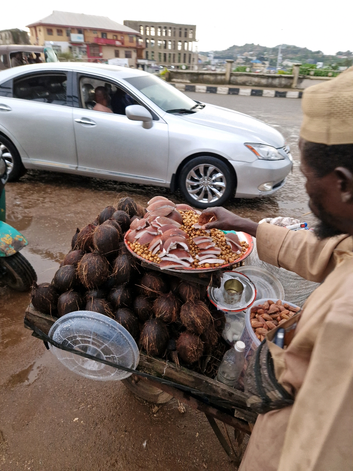 The image depicts a street scene in Jos, Nigeria, with a focus on a street vendor selling coconuts and nuts from a cart. A silver sedan is parked on the wet, paved road, with a person visible in the driver's seat. The vendor, a dark-skinned man wearing a light brown traditional outfit and a cap, is seen interacting with his merchandise. He is reaching into a bowl of small, light brown nuts. The cart is laden with whole coconuts, their husks intact, and a large, shallow bowl filled with sliced coconuts and more of the small nuts. In the background, several buildings are visible, including a multi-story structure with exposed concrete framework, suggesting ongoing construction. The overall environment appears to be an urban setting with a somewhat overcast sky, contributing to the wet appearance of the road.