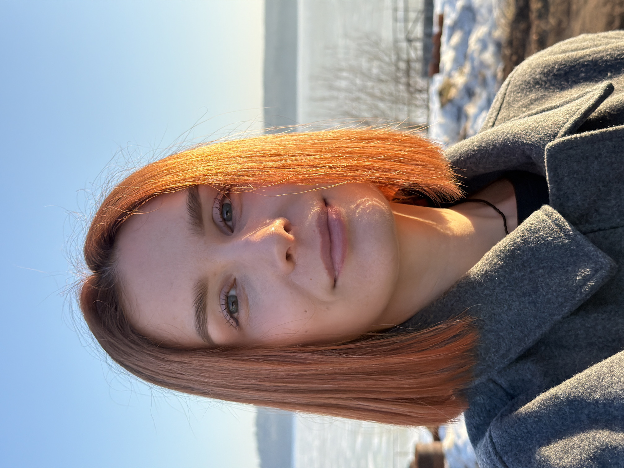 A young woman with short, reddish-orange hair is shown in a portrait-style image. She has fair skin, light eyes, and a neutral expression. She is wearing a dark-colored top and a grey wool coat. The background appears to be outdoors, featuring a body of water (possibly a lake or river) under a blue sky. Some bare trees and snow-covered ground are also visible, suggesting a winter or early spring setting. The image may have been taken near the city of Chaikovsky, Russia, due to the request to locate it there.