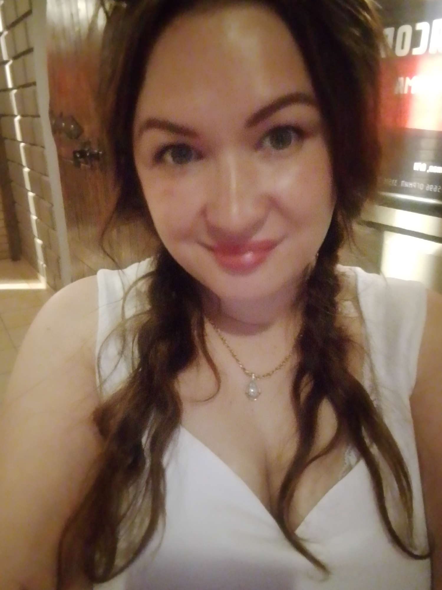 A selfie depicts a woman with fair skin, shoulder-length brown hair braided into two pigtails, and wearing a white v-neck top and a delicate gold necklace with a teardrop pendant. She is smiling softly at the camera.
In the background, partially visible, is a wall featuring a vertical row of textured tiles, suggesting an interior setting. Another, reddish, abstract sign is visible but too blurry to make out the words. The image's low lighting conditions give the scene a warm tone.
There is no information indicating the location is in Chaikovsky, Russia.