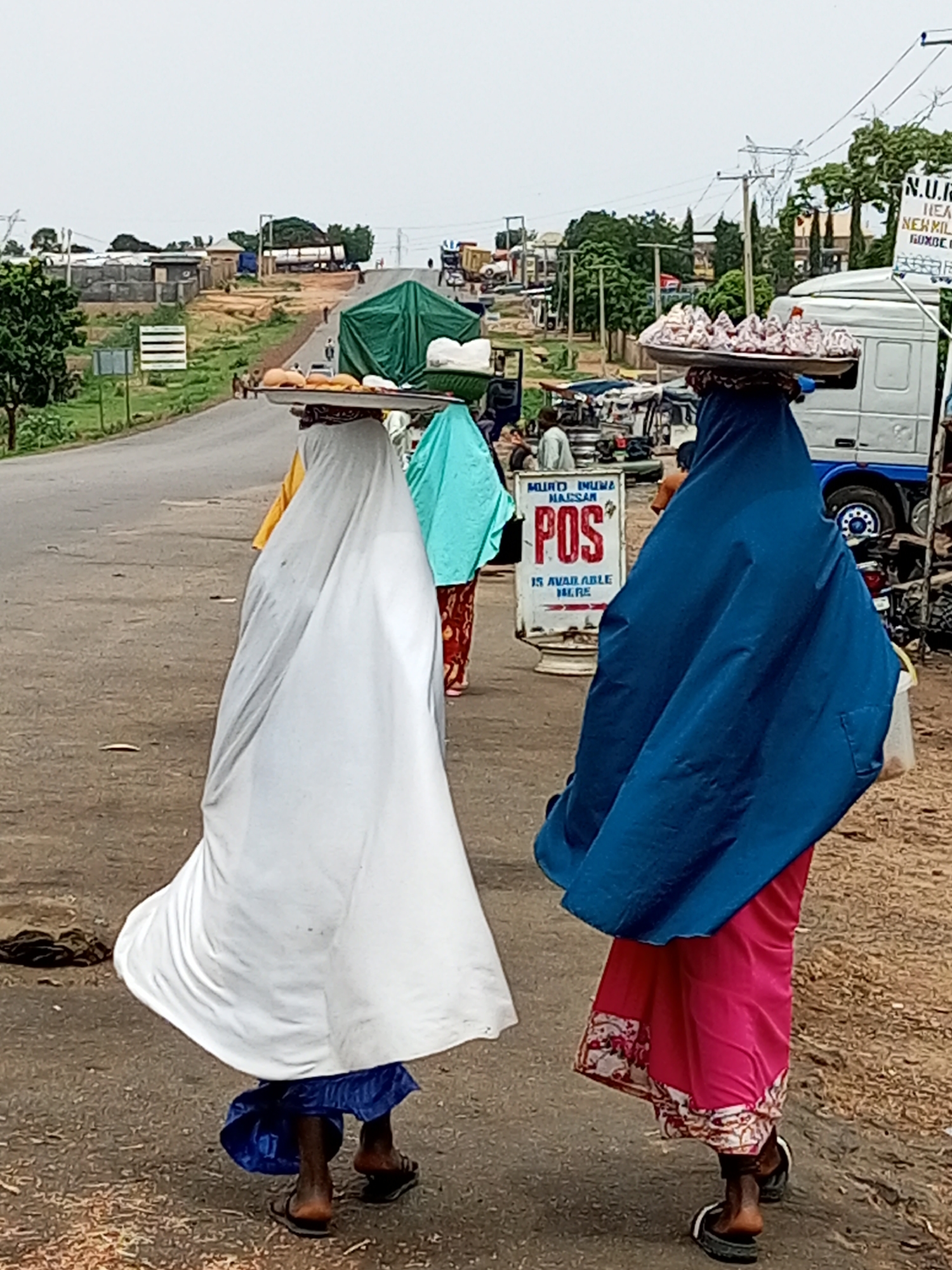 In the city of Gadao, Nigeria, two women are walking away from the camera along a paved road. The woman on the left is wearing a white hijab and a blue wrap around her waist and legs. She is carrying a tray of food balanced on her head, which appears to contain bread rolls and other items. The woman on the right is wearing a blue hijab and a pink skirt. She is also carrying a tray on her head, loaded with what look like pastries or sweets. Behind them, there are various vehicles, including a white truck, and market stalls. The environment suggests a bustling roadside market.