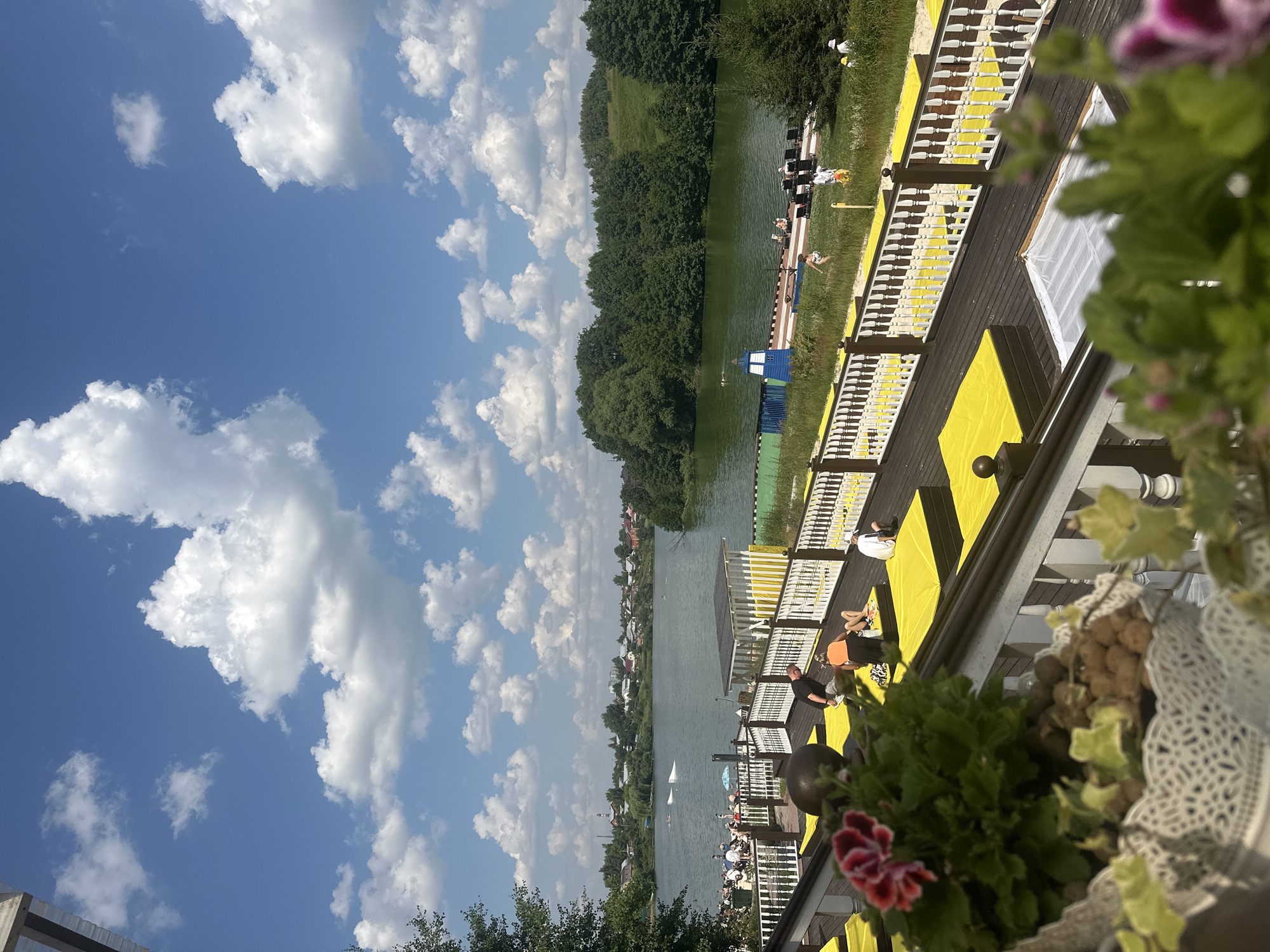 This media file depicts a sunny day scene by a body of water, likely a lake or river, at Dal'nie Prudishchi, Russia. The perspective is from a wooden deck with yellow and white accents, overlooking the water.
The primary subjects are people relaxing on the deck and in the water. On the deck, several individuals are seated or lying down on yellow mats, enjoying the sunshine. Some appear to be sunbathing or socializing. In the water, a few people are visible, possibly swimming or engaged in water activities. There are also small boats on the water.
The scene is set on a wooden pier or deck with a railing. Yellow mats are spread out on the deck, creating a vibrant visual element. Green grass borders the deck and slopes down towards the water. A line of trees forms the background on the opposite bank, with some houses visible in the distance, suggesting a semi-rural or recreational area.
The weather appears to be clear and warm, indicated by the bright sunshine and scattered white clouds against a deep blue sky. The time of day is likely midday or early afternoon, given the strong sunlight. There is no visible text in the image. The overall mood is one of leisure and relaxation on a summer day.