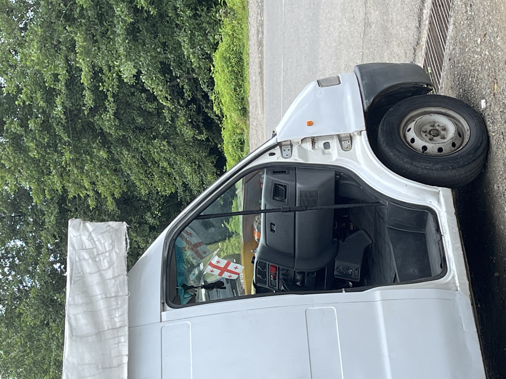 The image shows a white van parked on the side of a road in Ureki, Georgia. The van's driver's side door is open, revealing the interior dashboard and driver's seat. A small Georgian flag is hanging from the rearview mirror inside the van. The van is parked on a gravel surface next to a paved road, with a dense green forest in the background. The overall scene appears to be a roadside stop in a natural setting.