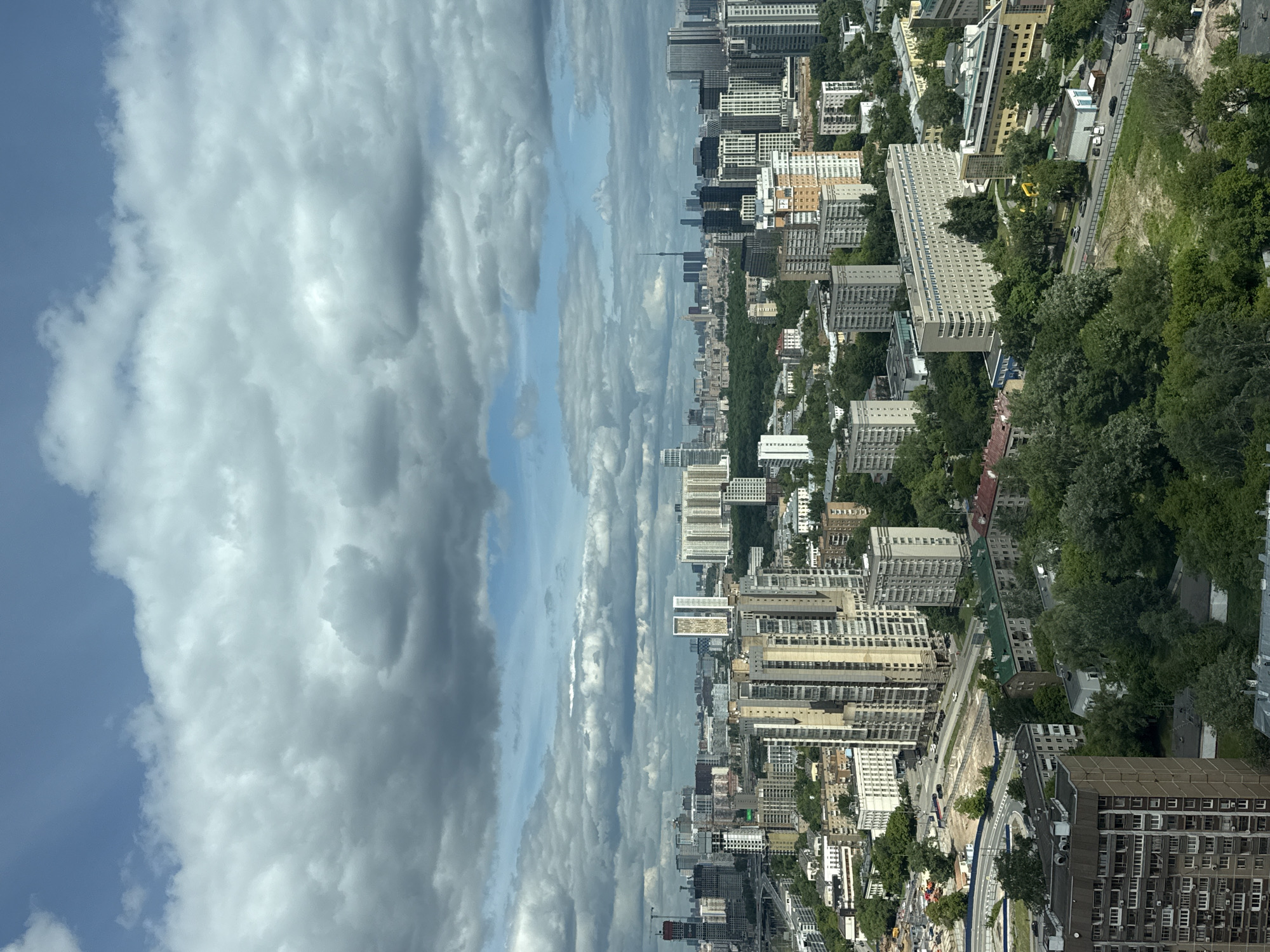 The image shows an aerial view of a city. The scene is split between a cloudy sky on the left and a cityscape on the right. The buildings are mostly residential, with some taller structures towards the center. Trees are interspersed among the buildings. Cars can be seen on the streets.
Based on visual features, including the architectural styles and landscape, the city is Khimki, Russia. The image appears to be taken from a high vantage point, such as from a tall building.