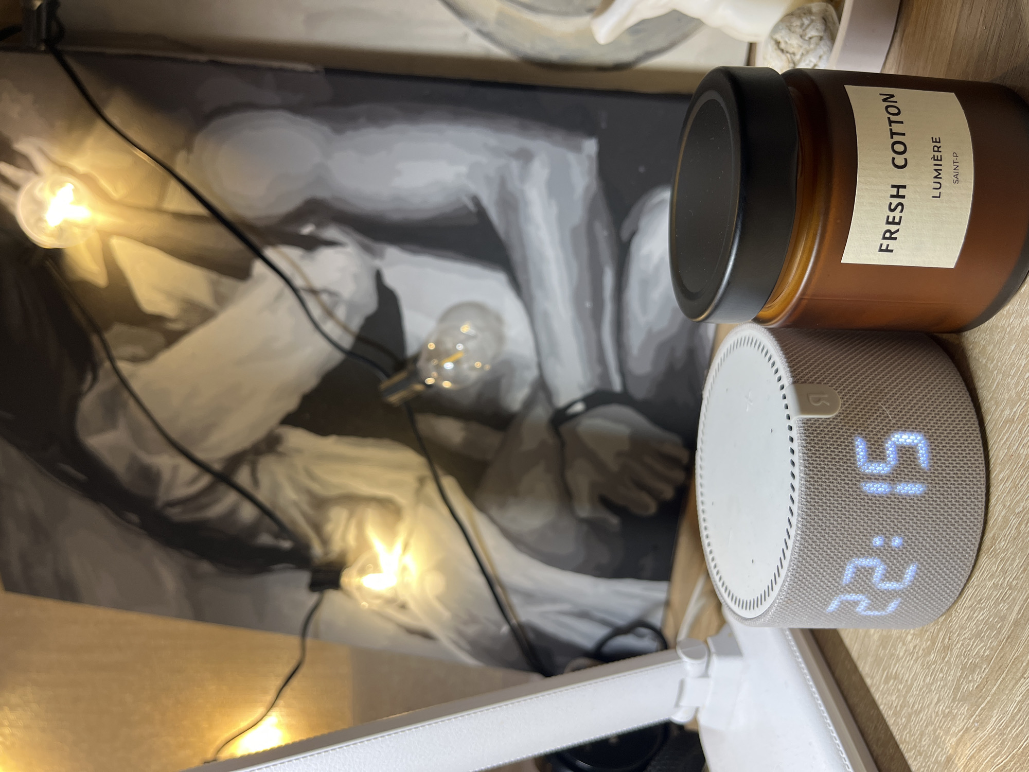 A brown glass candle jar with a black lid, labeled "Fresh Cotton Lumière Saint-P," sits next to a spherical smart speaker that displays "22:15." These are positioned on a wooden surface near a black and white drawing illuminated by string lights. A portion of a white statue is visible in the background. A white articulating desk lamp is also present. The image was taken indoors, with the camera positioned at an oblique angle. There is no evidence to suggest the location is Sochi, Russia.