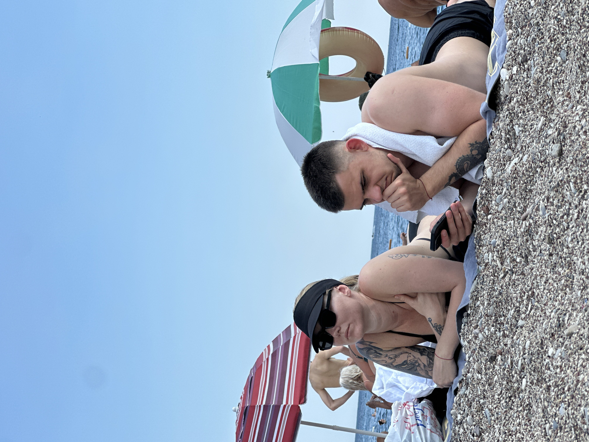 A man and a woman are lying on a pebble beach in Boreti, Montenegro. The woman, on the left, wears a black bikini top and a black visor. She has tattoos on her arms. The man, on the right, wears black swim trunks and has a white towel draped around his neck. He is looking at his phone. A red and white striped umbrella is behind the woman, and a green and white umbrella is behind the man. The sea is visible in the background.