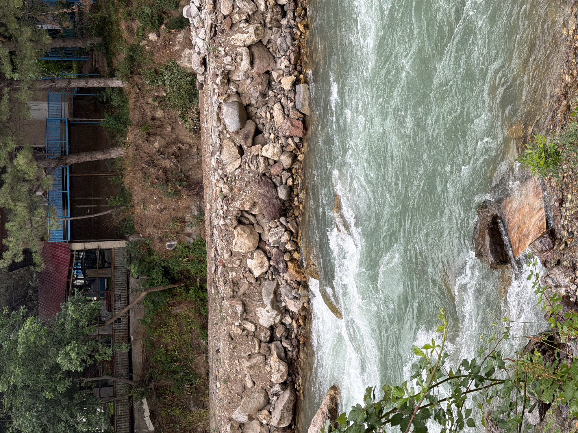 The image depicts a fast-flowing, turquoise river in Khumsan, Uzbekistan. The riverbanks are lined with a mixture of rocks and gravel. Above the river, on the embankment, is a structure that appears to be a building with a red metal roof and blue railings. Trees and vegetation are present on the embankment and surrounding the building. The scene is naturalistic, with the river being the dominant feature. There are no people or animals visible in the image. The lighting suggests it is daytime.