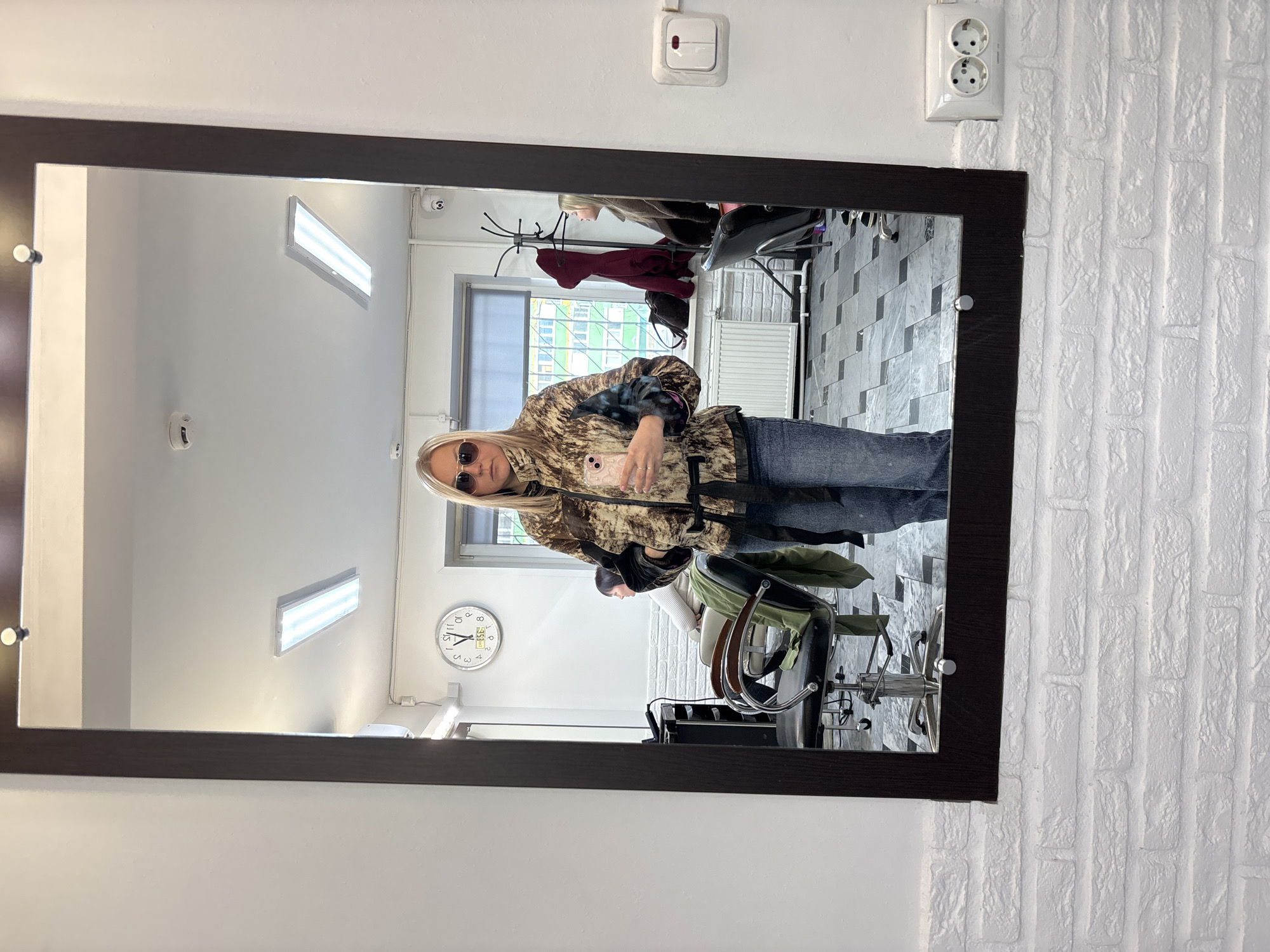The image depicts a mirror reflection of a woman holding a small dog in what appears to be a beauty salon or hairdressing studio in Izhevsk, Russia.
The woman, wearing sunglasses and a patterned jacket with jeans, looks towards the mirror while holding the dog. In the reflection, a second person is partially visible sitting in a salon chair with green fabric. The salon appears to have white brick-patterned walls, grey tile flooring, fluorescent lighting, and standard hairdressing equipment. A clock on the wall shows the time is approximately 8:03. In the upper left of the mirror, a clothes rack and an open window are also visible. The mirror is a rectangular, dark-framed object mounted on the wall next to a double electrical outlet and a single light switch.