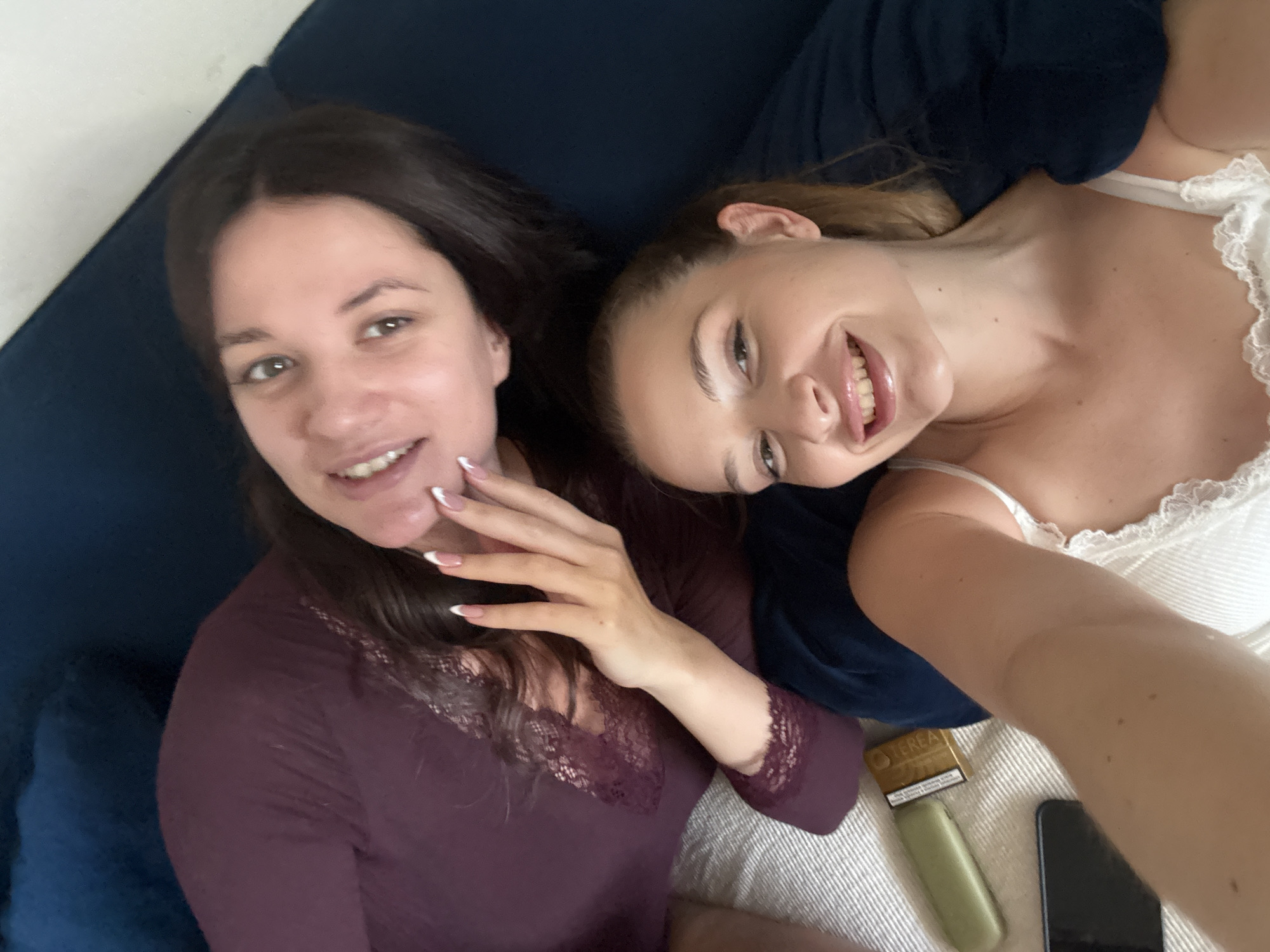 Two women are lying on a blue couch and a light-colored surface. The woman on the left has dark hair and is wearing a dark-colored shirt. She is touching her face with her hand. The woman on the right has light brown hair and is wearing a white lace-trimmed top. A mobile phone and a vape pen are visible on the light-colored surface. The location is Belgrade, Serbia.
