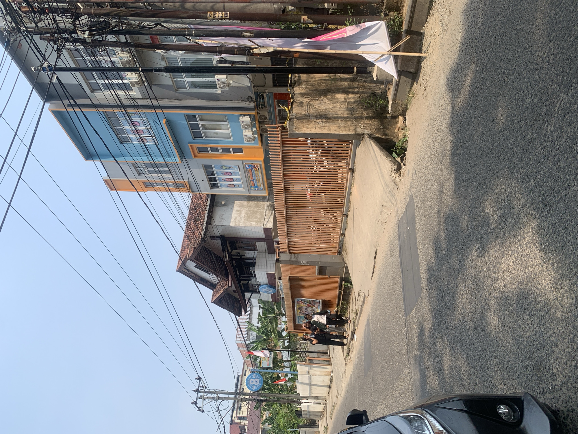 The image captures a street scene in Palembang, Indonesia, with a focus on the architecture and urban environment. In the foreground, the left side of a dark-colored car is visible, parked on a paved road. The road surface shows signs of wear and tear with patches of darker asphalt. To the right of the road, a sidewalk and a concrete wall lead up to a building complex.
The building complex features a mix of architectural styles. On the left, a traditional-looking house with a reddish-tiled roof and wooden balconies stands behind a white fence. Adjacent to this, a modern, multi-story building displays a vibrant facade with sections painted in blue and orange, contrasting with grey walls. This building has numerous windows and visible air conditioning units.
A prominent feature of the scene is a tall wooden fence with vertical slats, suggesting a boundary or entrance to a property. Three individuals, two women in dark clothing and one in a lighter outfit, are walking on the sidewalk near the fence.
The environment is characterized by overhead power lines and utility poles, indicating the presence of electricity and telecommunication infrastructure. A white flag with pink accents is propped up on a bamboo stand near the wall. The sky is clear and blue, suggesting fair weather. The overall impression is a bustling urban street with a blend of older and newer structures, typical of a city in Indonesia.