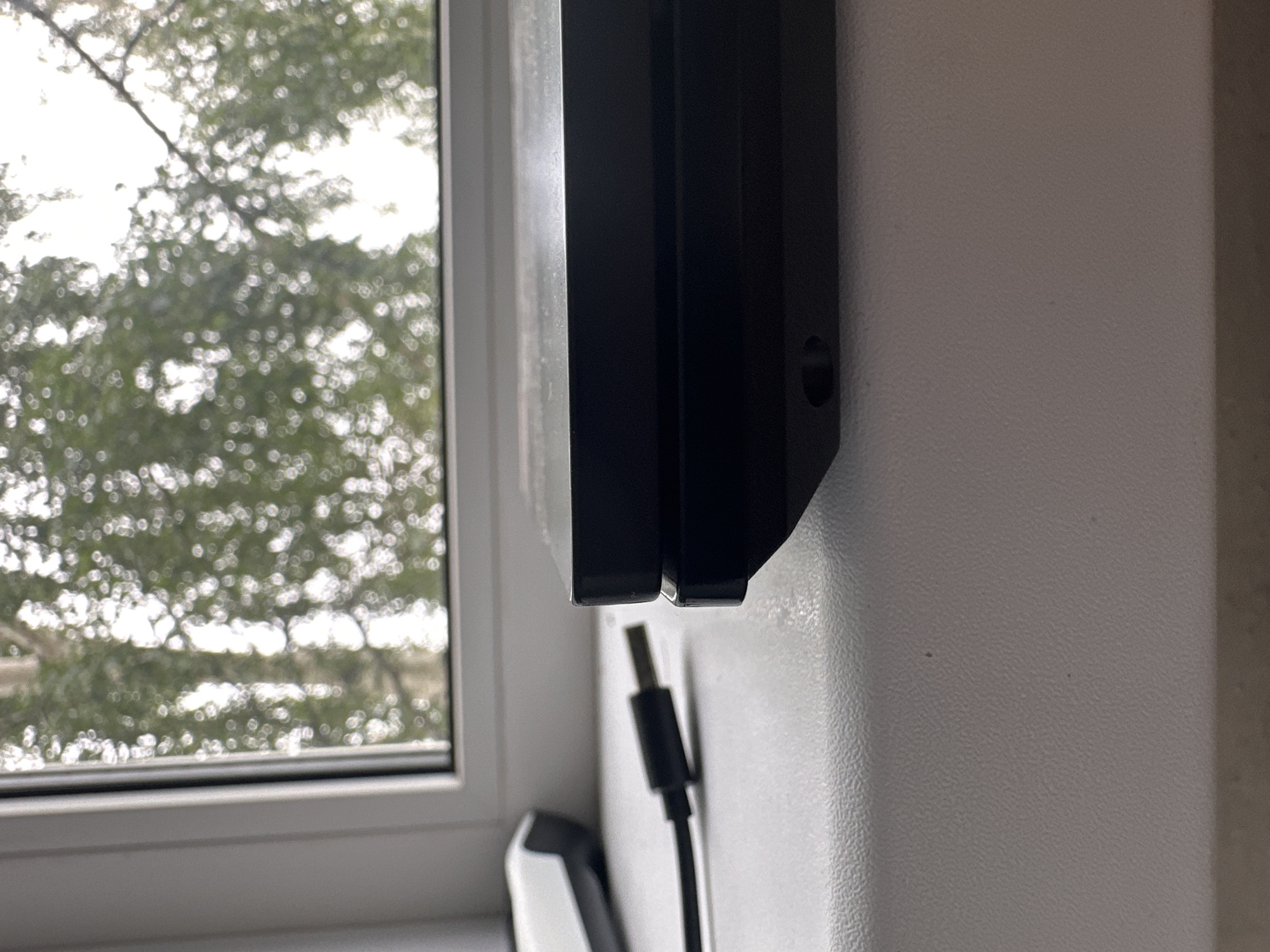 The image shows a close-up view of a black electronic device, likely a game console or external hard drive, placed on a white windowsill. A USB cable with a silver connector is plugged into the side of the device and extends out of frame. In the background, a window displays a blurry view of green foliage and bright sky, suggesting an outdoor scene. The overall environment appears to be indoors, possibly a room with a window overlooking trees. The image is taken from a low angle, focusing on the electronic device and the windowsill. There are no people visible in the image. The specific location in Plesetsk, Russia cannot be determined from the visual information provided.