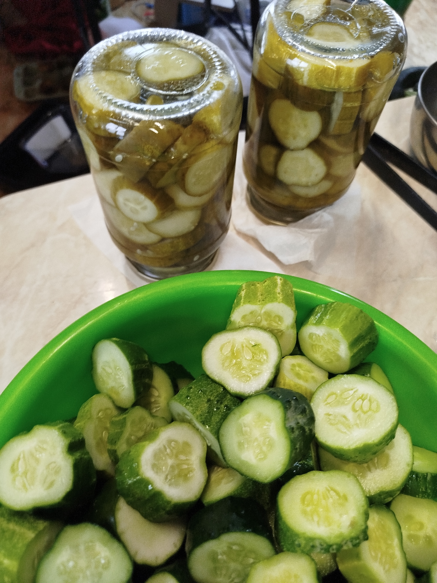 In Zavodoukovsk, Russia, on a countertop, are two jars of pickled cucumbers. In the foreground, a green plastic bowl overflows with sliced cucumbers. The jars, filled with sliced pickles submerged in brine, are positioned behind the bowl. The surface they rest on appears to be a marble-patterned countertop. The scene is captured from a slightly elevated perspective, focusing on the food preparation.