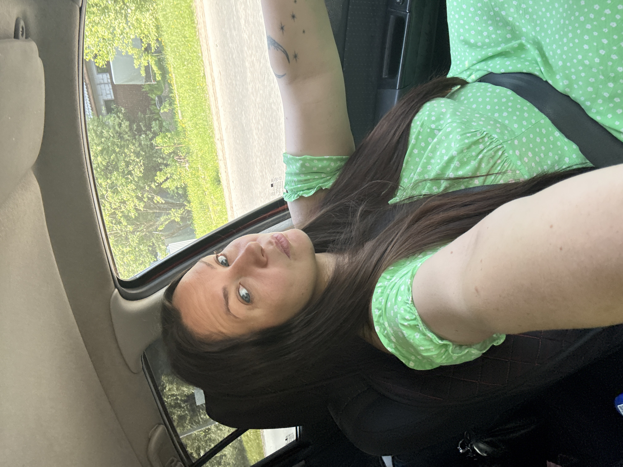 A woman is sitting in a car, wearing a light green, white-dotted dress. She is wearing her seatbelt. Her right arm is raised to the right of the frame. Her left arm is above her head. A crescent moon tattoo is visible on her left arm. Outside of the car, trees and a building can be seen. The location is the city of Chaikovsky, Russia.