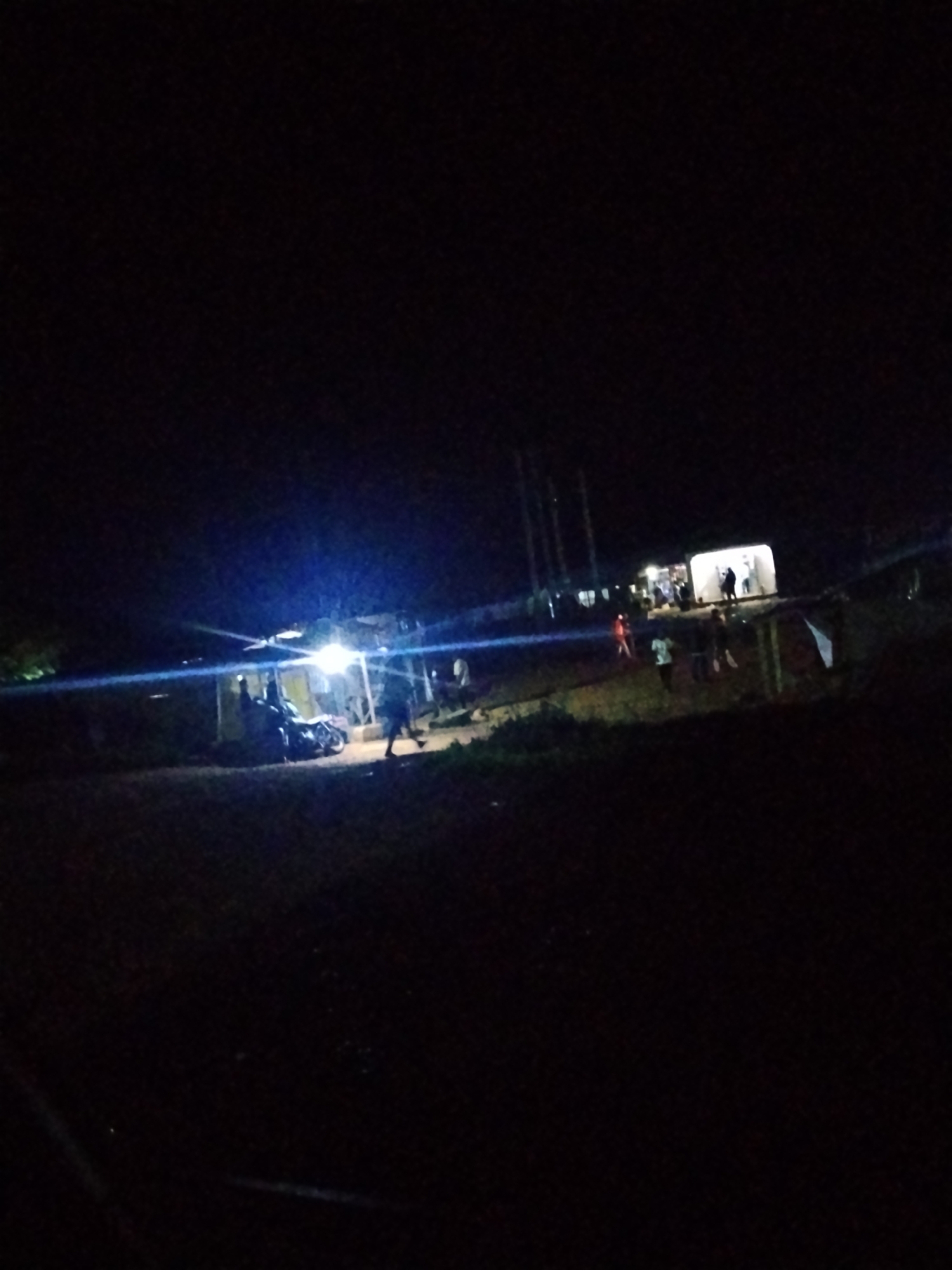 At night in Durumi, Nigeria, several people are gathered in a dimly lit area near a building that appears to be a shop or roadside stall. A motorcycle is parked to the left of the building, and a few individuals are standing around it. In front of the building, more people are visible, some walking and others conversing. The scene is illuminated by a bright light source originating from the building, casting shadows and creating a somewhat hazy atmosphere. The background is dark, suggesting it is open country or a less developed part of the city.