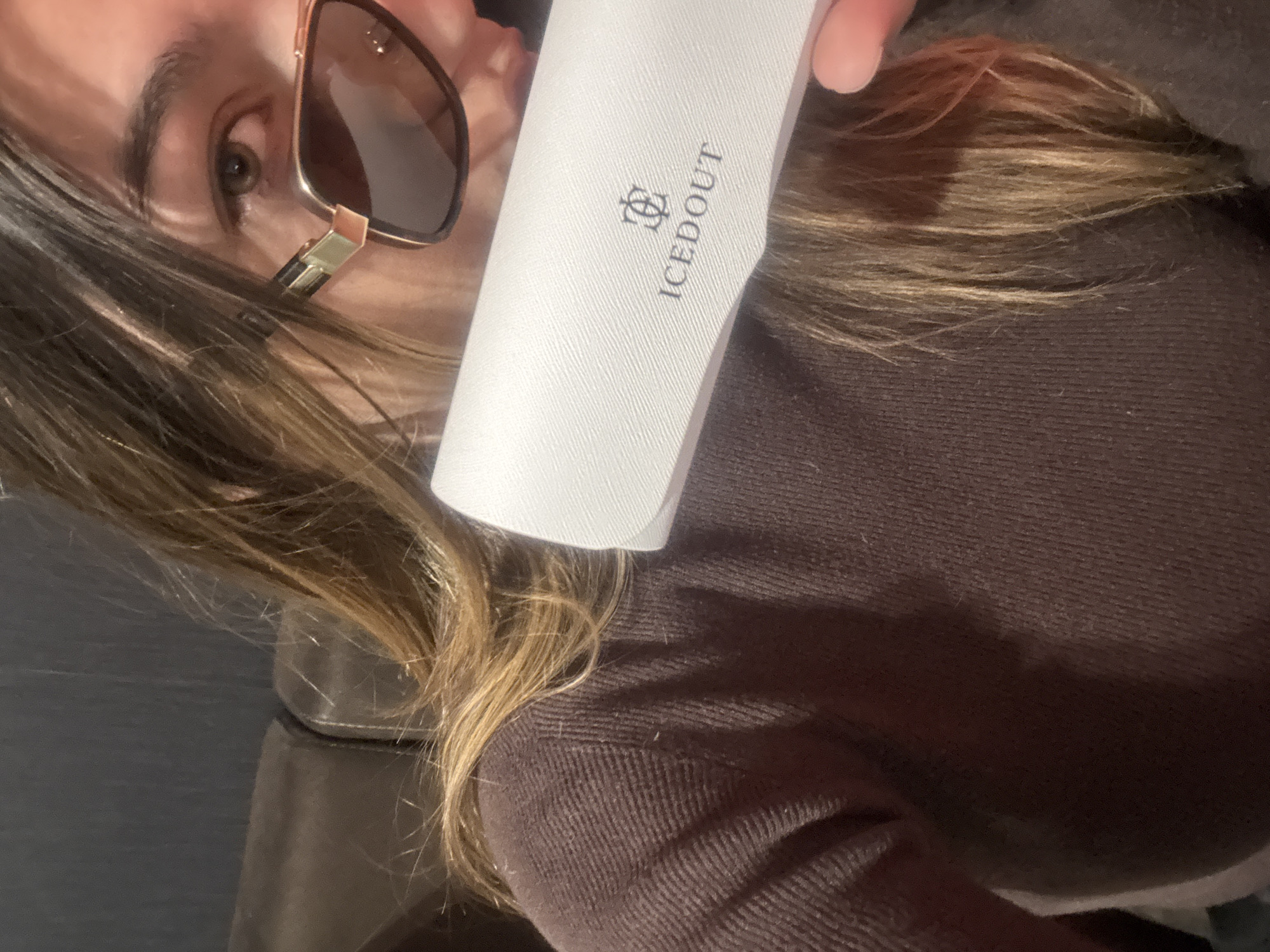 A person with brown and blonde hair is holding a white sunglasses case in front of them. The case has the logo "ICEDOUT" printed on it. The person is wearing sunglasses with a gold frame. The background is a brown sweater and an undefined dark surface. The location and city are unknown.