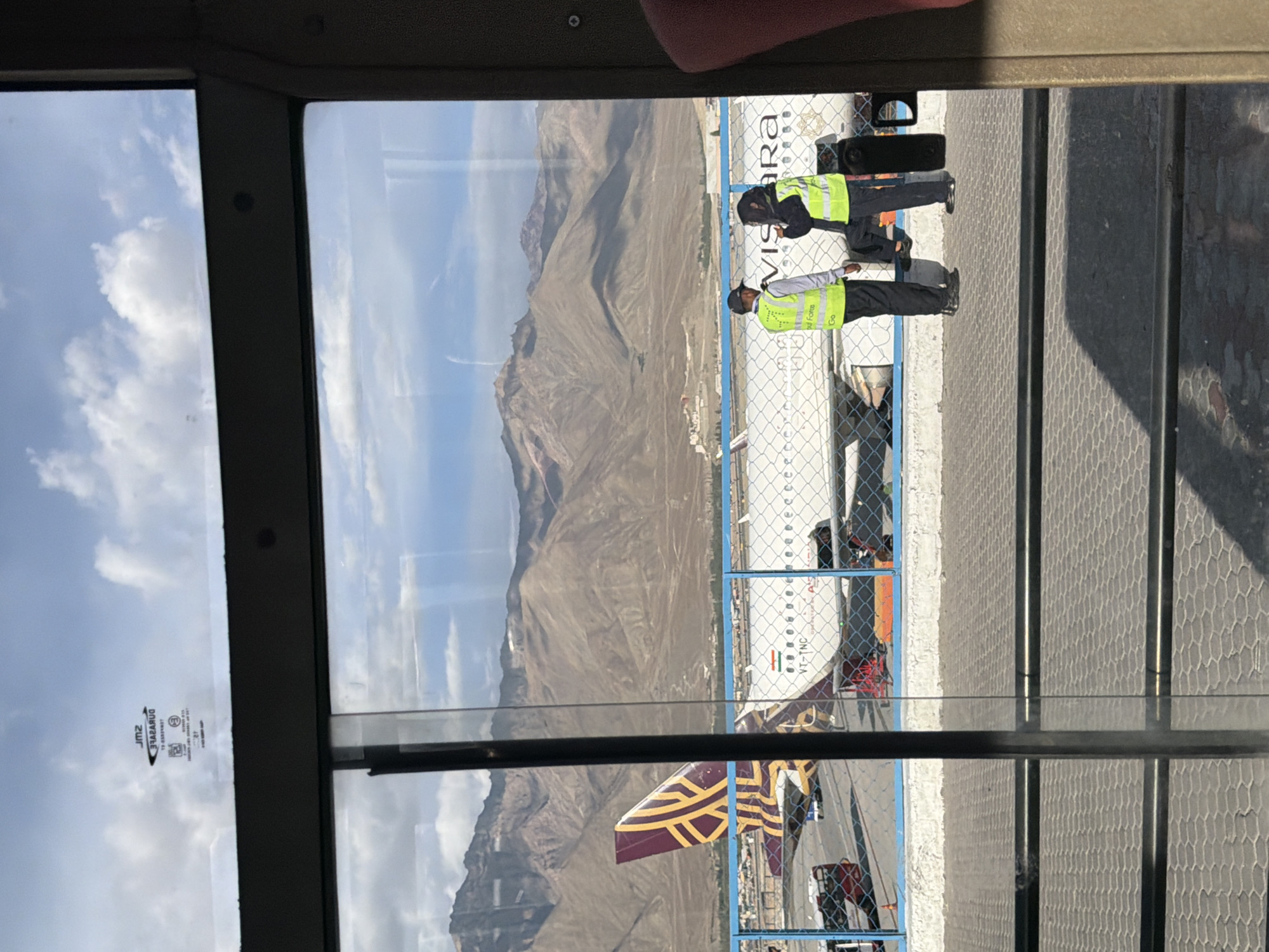 An exterior shot of an airport tarmac in Leh, India, is framed by a window. The tarmac is gray and paved with hexagonal tiles. A white fence with blue posts separates the tarmac from a waiting area. In the background, a large passenger airplane with a distinctive burgundy and gold tail is parked. The plane's fuselage is white, with the text "VISARA" prominently displayed. A small Indian flag is also visible on the side of the plane. Two individuals, dressed in bright yellow safety vests and dark pants, stand in conversation near the plane. One of them is wearing a dark baseball cap. In the far background, rugged, dry, mountainous terrain under a partly cloudy sky dominates the landscape. A few vehicles are also visible on the tarmac, including a red bus. The scene is illuminated by natural daylight.