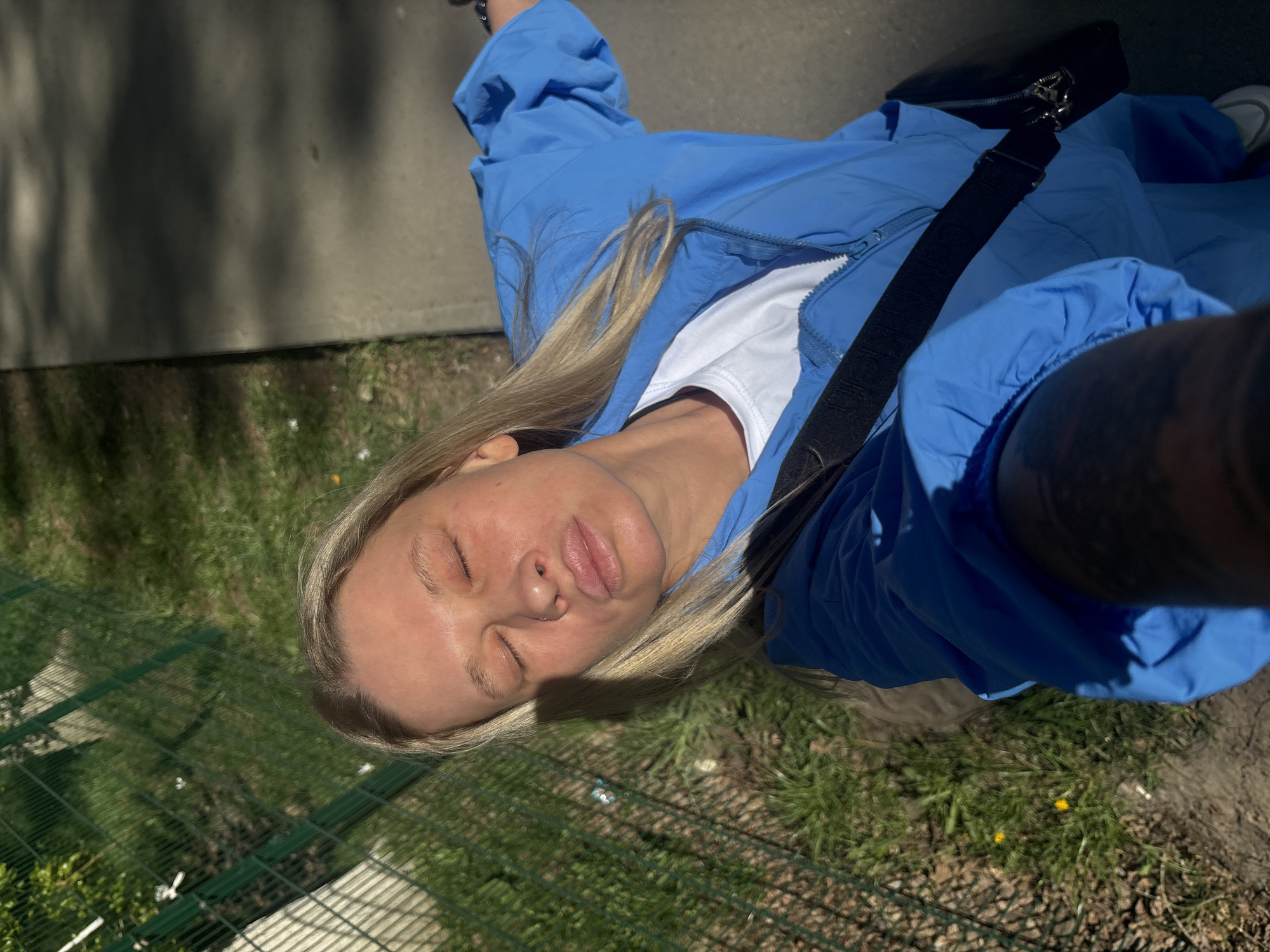 A woman with fair skin, blonde hair, and closed eyes is positioned at an angle in an outdoor setting. She wears a blue jacket over a white shirt and a black strap is visible over her shoulder. Part of her arm, with a tattoo, is in the foreground. A grassy area is visible below, along with a concrete surface, possibly a sidewalk. It is difficult to confirm the location as Perm, Russia, without additional context or visual clues specific to the city.