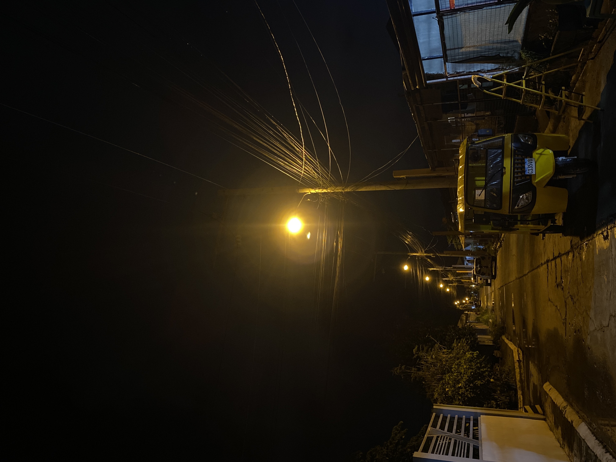 The image depicts a street scene at night in what appears to be a residential area of Davao City, Philippines. A yellow motorized tricycle is parked on the side of a narrow, paved street that is wet, possibly from recent rain, with puddles reflecting the yellow glow of streetlights. The street is lined with various structures, some appearing to be small houses or shops with corrugated metal roofs. A yellow stepladder is propped against one of the buildings. Overhead, a complex network of electrical wires crisscrosses the dark sky, illuminated by a bright streetlamp positioned on a utility pole. Trees and foliage are visible along the sides of the street, adding to the urban yet somewhat natural setting. The overall impression is one of a quiet, nocturnal neighborhood scene in the Philippines.
