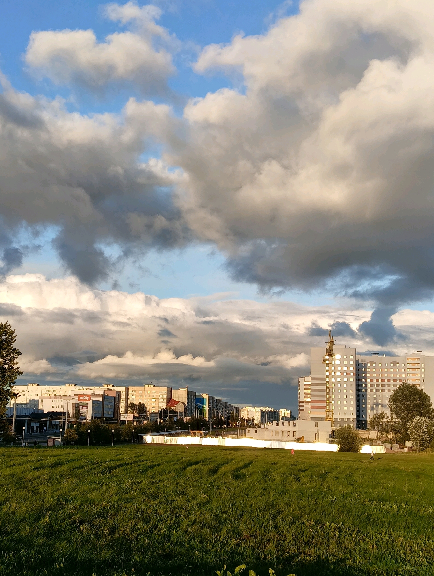 This image captures a view of the Peryastrowsky District in Hrodna, Belarus, during what appears to be late afternoon or early evening. The foreground is dominated by a lush, green grassy field, indicating a park or undeveloped area. Beyond the field, a cityscape unfolds with several multi-story apartment buildings. The architecture suggests a post-Soviet era construction style, with a mix of neutral and colorful facades. In the center-right, a prominent, tall residential building stands out, its windows reflecting the warm light of the setting sun. A construction crane is visible on top of this building, indicating ongoing development or renovation. To the left, a line of lower-rise buildings forms the urban skyline, some of which appear to be commercial establishments based on visible signage. A lone tree stands on the far left edge of the frame, adding a touch of nature to the urban scene. The sky is a dramatic display of clouds, with a mix of dark, ominous storm clouds and bright, sunlit patches, suggesting variable weather conditions. Two figures, likely pedestrians, are present in the middle ground, walking across the grassy area, suggesting a public space that is utilized by residents.