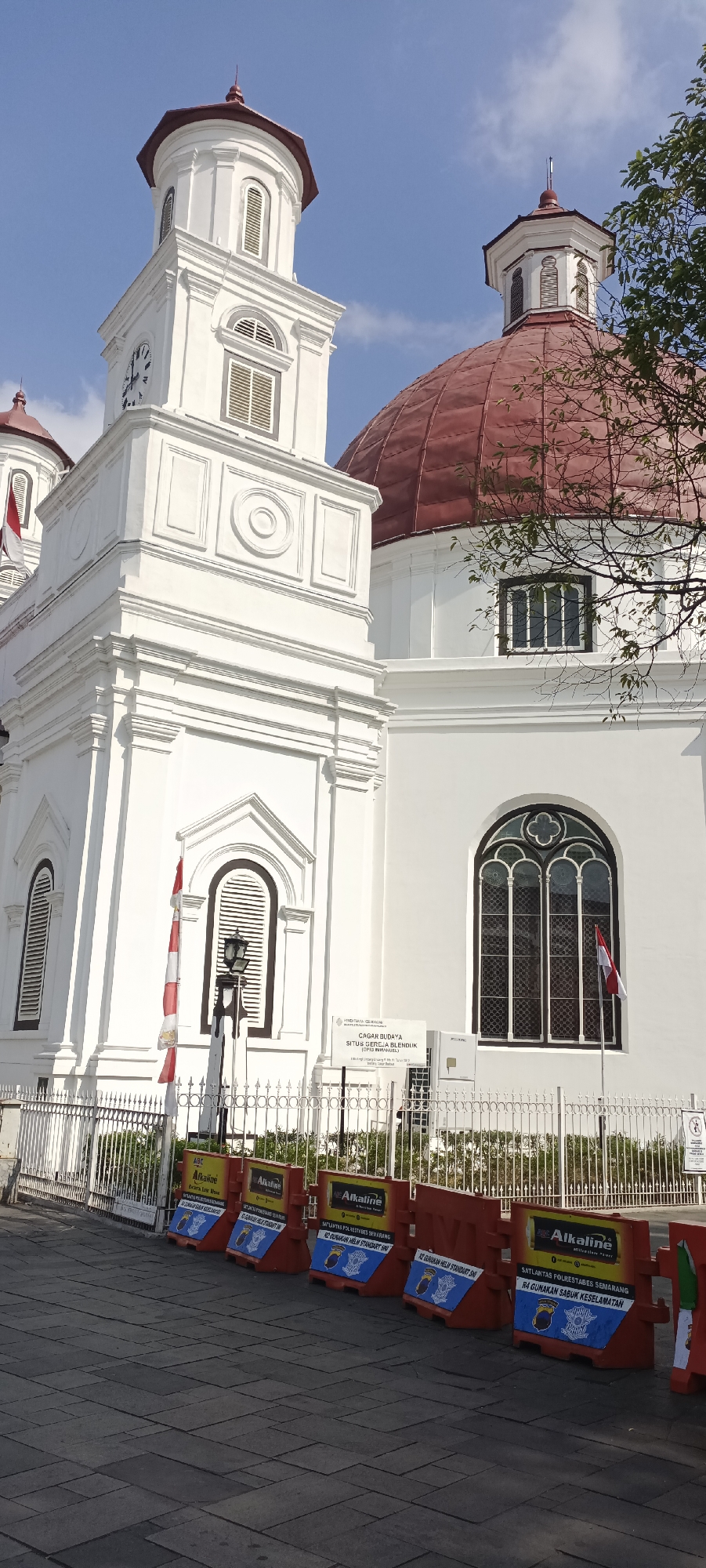 The image shows the Blenduk Church, a historical landmark in Semarang, Indonesia. The church, painted white, features a prominent red dome and a clock tower. In front of the church, there is a white metal fence. Several red and yellow traffic barriers are placed along the sidewalk. The barriers have text and logos on them, including the word "Alkaline" and what appear to be police insignias. There are also two Indonesian flags visible, one red and white flag on a flagpole near the church entrance, and another smaller red and white flag attached to a barrier. The sky is clear and blue, suggesting a sunny day. A tree branch with leaves is visible in the upper right corner of the image, partially obscuring the church dome. The ground in the foreground is paved with dark gray square tiles.