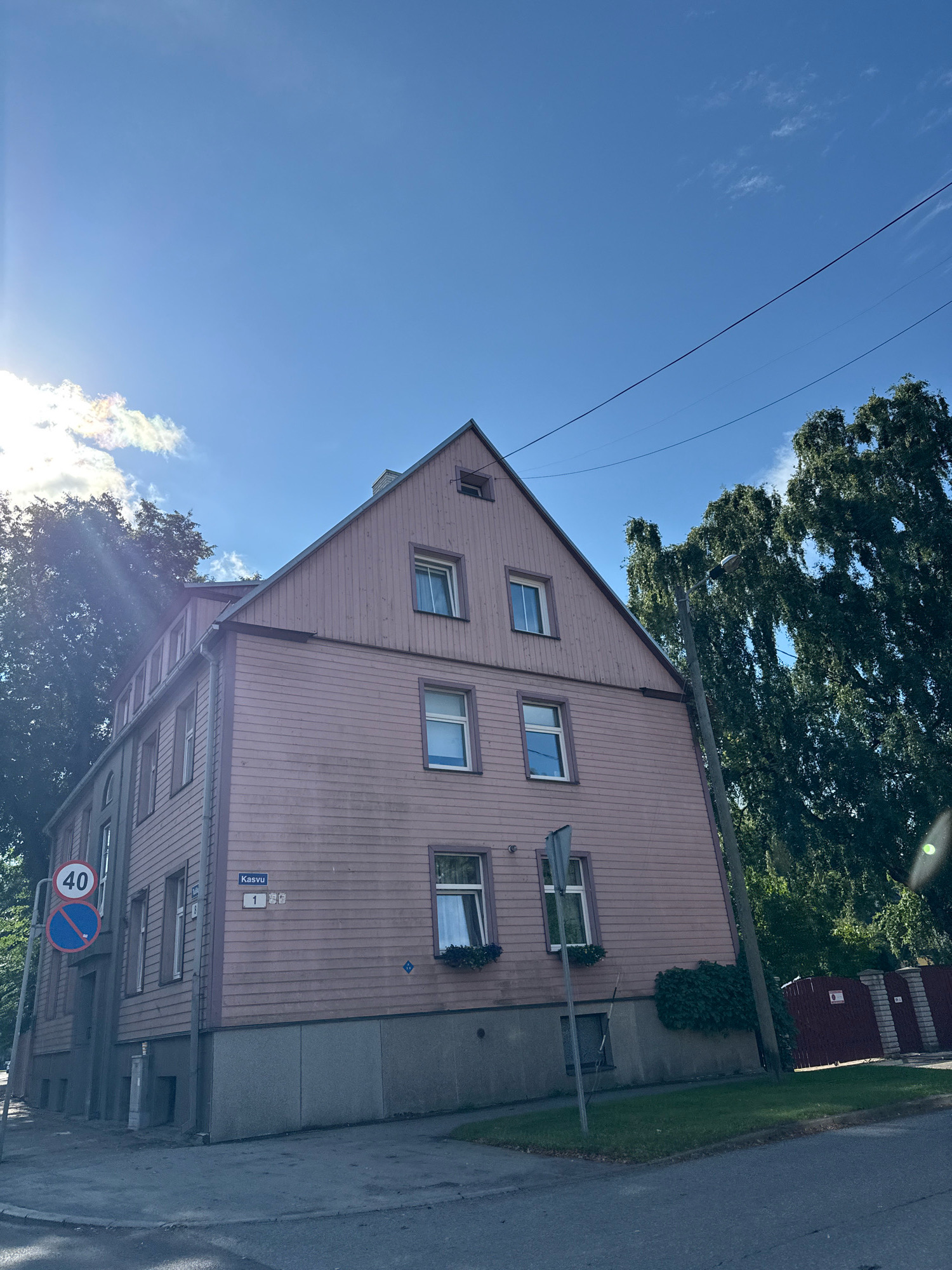 The image shows a three-story wooden house painted pink, located on Kasvu Street in Tallinn, Estonia. The house has a steeply pitched roof and several windows on each level. A speed limit sign for 40 kilometers per hour is visible on the left side of the image, along with a blue sign indicating no stopping. A street lamp is positioned in front of the house, and a large, leafy tree partially obscures the right side of the building. The sky is blue with some scattered clouds, and the sun is shining brightly, casting a lens flare in the upper right corner of the image. The ground in front of the house is a paved sidewalk and a street. The overall scene depicts a residential area on a sunny day.