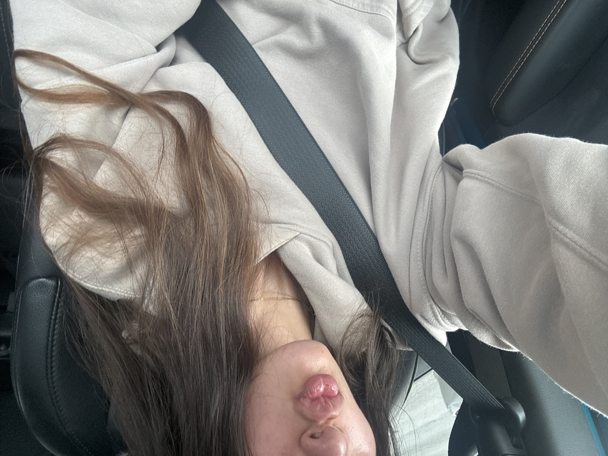 A young woman with long, brown hair and light skin is sitting in the driver's seat of a car, wearing a light-colored hooded sweatshirt and a thin gold necklace. Her lips are pursed in a kiss. A black seatbelt is fastened across her chest. The interior of the car appears to be black leather. The background is blurry, but suggests an outdoor environment. The image was taken from the woman's perspective.