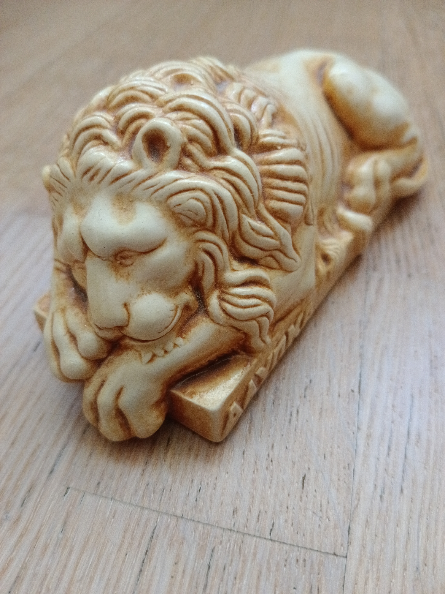 The image shows a close-up, high-angle shot of a light-colored, carved figurine of a lion resting on its paws. The lion is positioned diagonally, with its head facing left and its body extending towards the right of the frame. The figurine has detailed features, including a mane, eyes, nose, and paws. The surface beneath the figurine appears to be a light-colored wooden floor with visible grain. There is no indication of the city of Severnyi, Russia, or any specific environmental context beyond the floor.