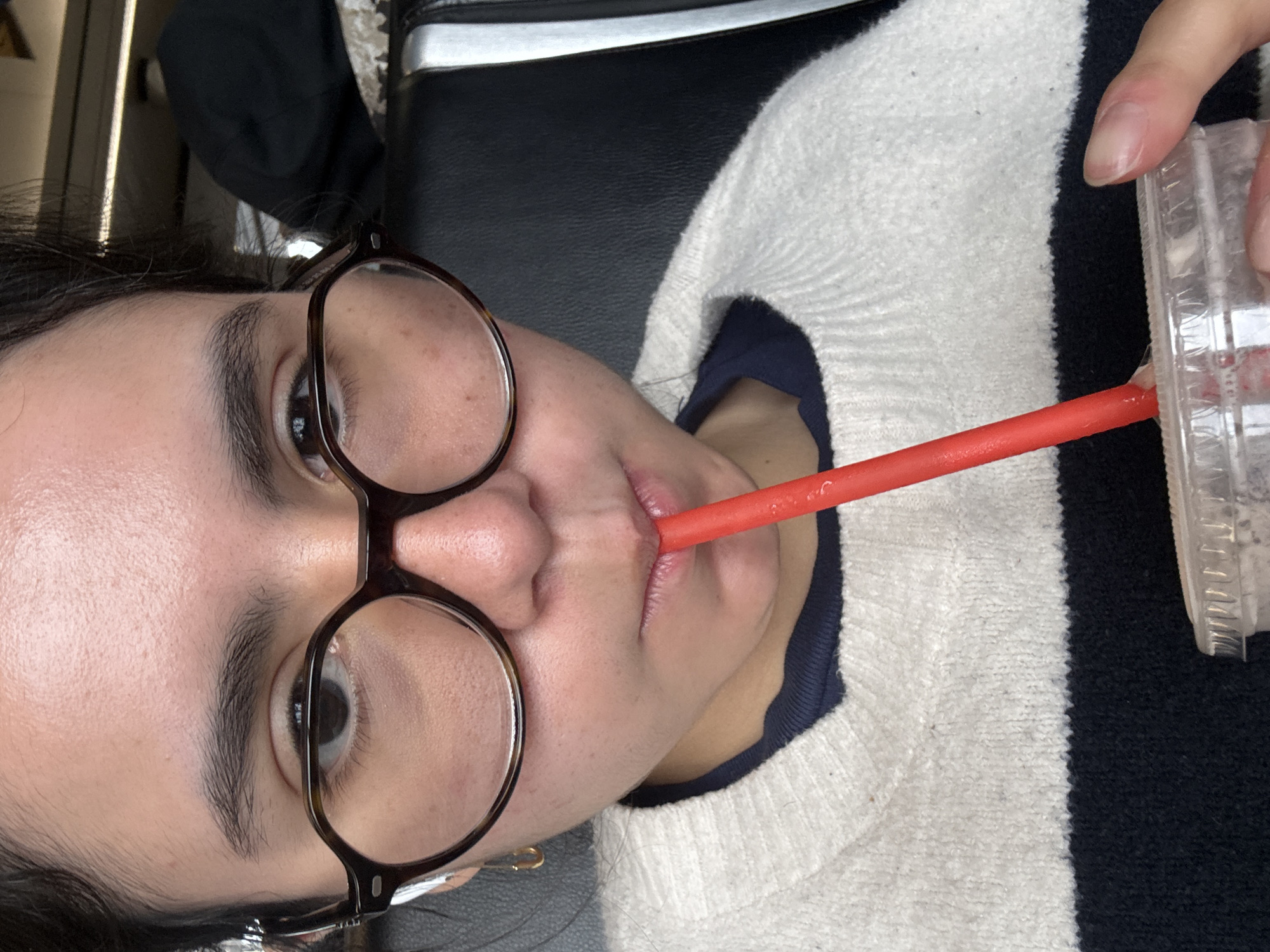 A person with dark hair and glasses is drinking from a clear plastic cup with a red straw. The person is wearing a striped sweater with white and black stripes, with a visible blue undershirt. The location is unspecified, and there is no visual information to suggest Chicago, United States of America.