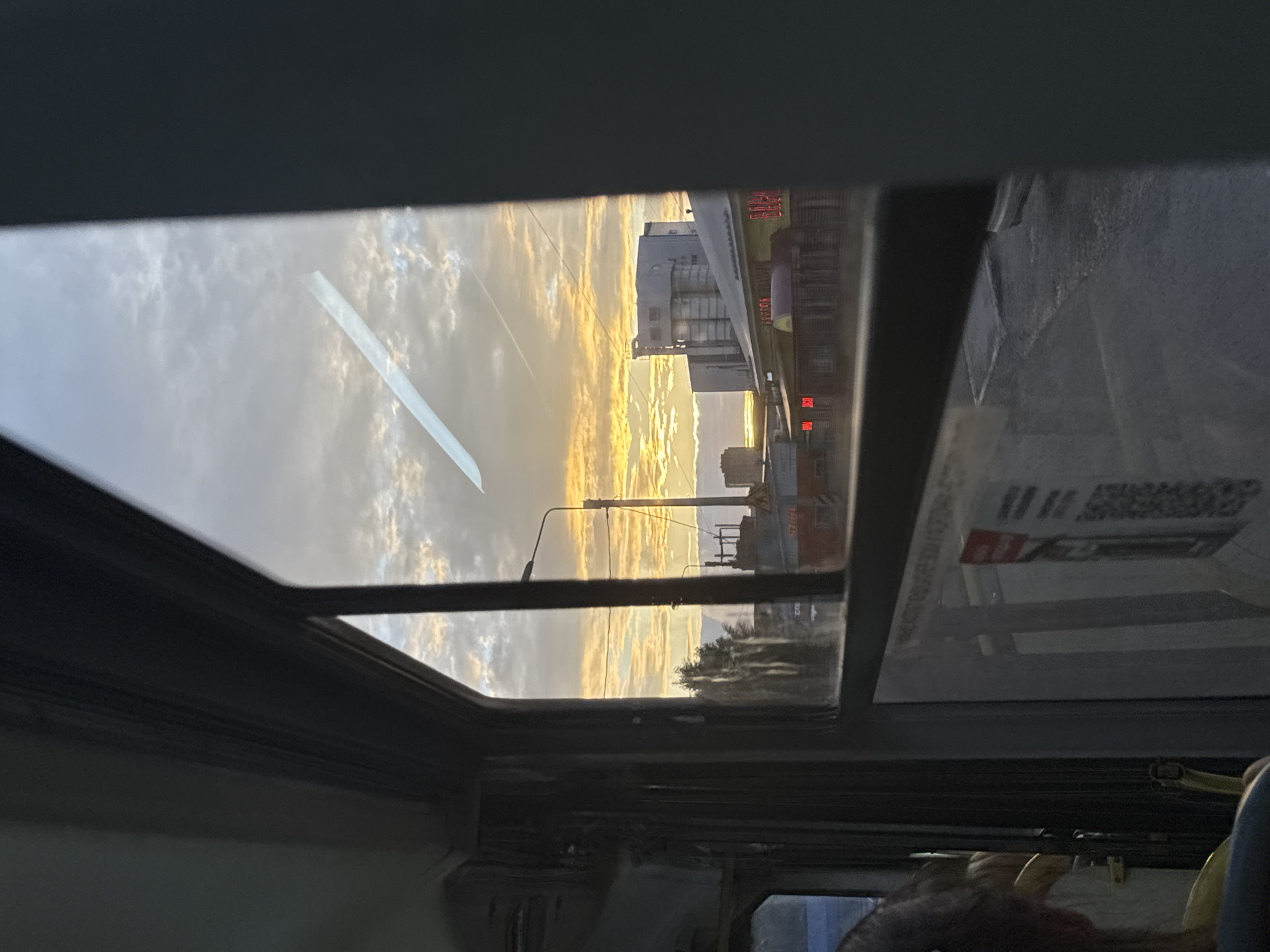 A view from inside a public transit vehicle, likely a bus or trolleybus, through a window overlooking an urban landscape during either sunset or sunrise. The sky is filled with scattered clouds, predominantly illuminated with warm golden-orange and yellow light, fading to lighter blue-grey tones towards the top. A faint, linear contrail is visible diagonally across the upper portion of the sky.
The vehicle's dark window frame, ceiling, and interior structures are visible, including a dark vertical pole on the left. A reflection of a bright, elongated object, possibly an interior light fixture, crosses the upper part of the window diagonally. In the lower left, the back of a passenger's head with light-colored hair and a portion of a yellow garment are visible. Several stickers, including one featuring a QR code and a red informational placard, are affixed to the lower section of the window glass.
Outside, a street scene unfolds. A tall street lamp pole stands vertically to the left of the central view. In the midground, a prominent yellow building with a red roof is visible, featuring multiple bright red neon signs in Cyrillic script. The most clearly legible sign spells "БАНЯ №13" (Bathhouse No. 13). Behind this yellow building, a large, multi-story grey industrial-style building with vertical structural elements is discernible, consistent with facilities like the Perm Flour Mill. Other smaller, less distinct buildings, utility wires, and some trees are scattered throughout the background. The ground visible beyond the vehicle is dark asphalt. A partial view of another vehicle is present on the far right side of the street. Based on the "Баня №13" landmark and Cyrillic script, the location is identified as Perm, Russia, specifically near Revolyutsii Street, 13.