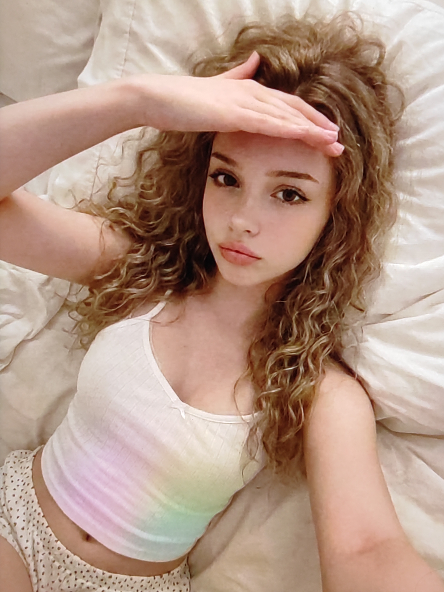 This media file features a close-up, overhead shot of a young woman lying in bed. She has curly, light brown hair and is looking up towards the camera with her right hand resting on her forehead. Her eyes are open and she appears to be looking directly at the viewer. She is wearing a white tank top with a rainbow tie-dye pattern and white polka dot shorts. The background consists of white bedding, suggesting an indoor setting, likely a bedroom. The lighting is soft and appears to be natural, possibly indicating daylight. The woman's expression is somewhat neutral, with a hint of curiosity. There is no visible text in the image. The scene is intimate and personal, focusing on the individual.