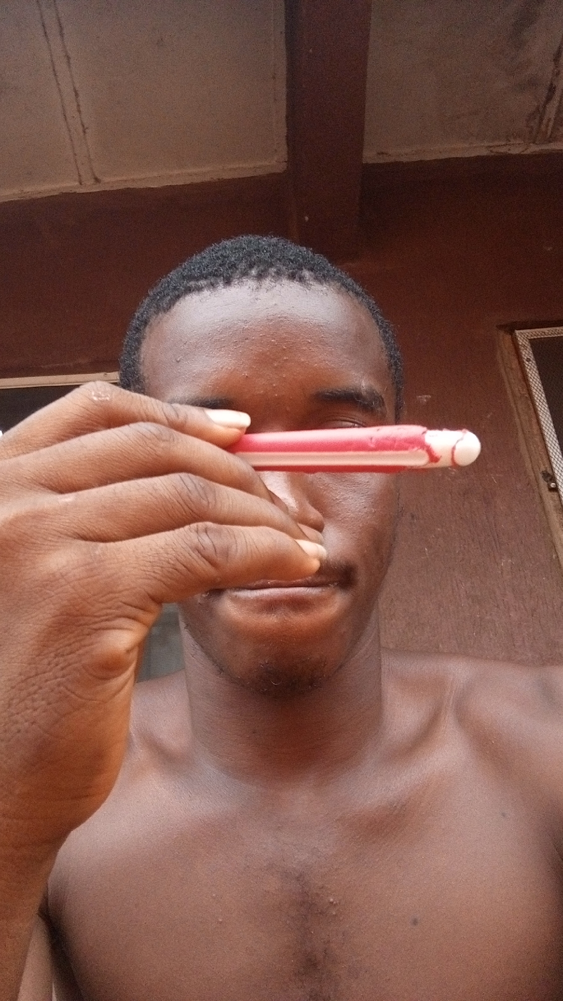 A shirtless dark-skinned man holds a red and white object horizontally across his right eye. The background consists of a weathered red-brown wall and a partially visible window. The location is implied to be Onitsha, Nigeria, but this detail is not visually verifiable from the image.