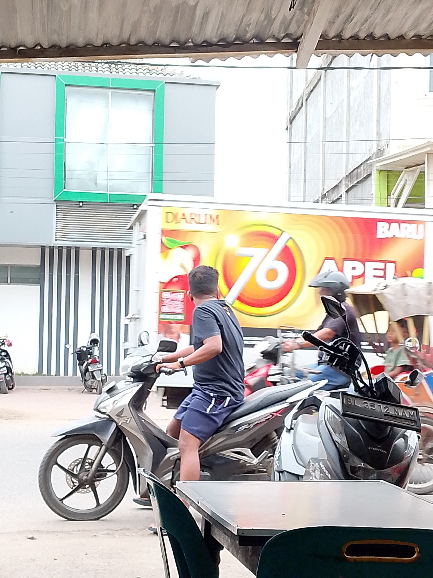 This media file depicts a street scene in Lhokseumawe, Indonesia. The central focus is on a man riding a modern, silver and black motorcycle. He is wearing a dark t-shirt and blue shorts, with a dark helmet covering his head. The motorcycle has a visible license plate reading "BL 3512 NAM." Another motorcycle, black and white, is parked behind him.
In the background, a large white truck is parked, featuring a colorful advertisement with bold yellow and red hues. Prominently displayed on the truck is the number "76" in a circular design, and text that reads "DIARUM" and "BARU APEL." To the left of the truck, a building with gray siding and a bright green-bordered window is visible, along with a row of vertical black and white striped columns. Further to the right, a glimpse of another building and possibly a market stall can be seen, with a child in a green shirt in the distance.
The overall impression is of a sunny day in an urban or semi-urban environment in Indonesia. The activity appears to be the movement of vehicles and people on a street, possibly near a commercial area given the truck advertisement and the presence of buildings. The scene is lively, suggesting typical daily activity.