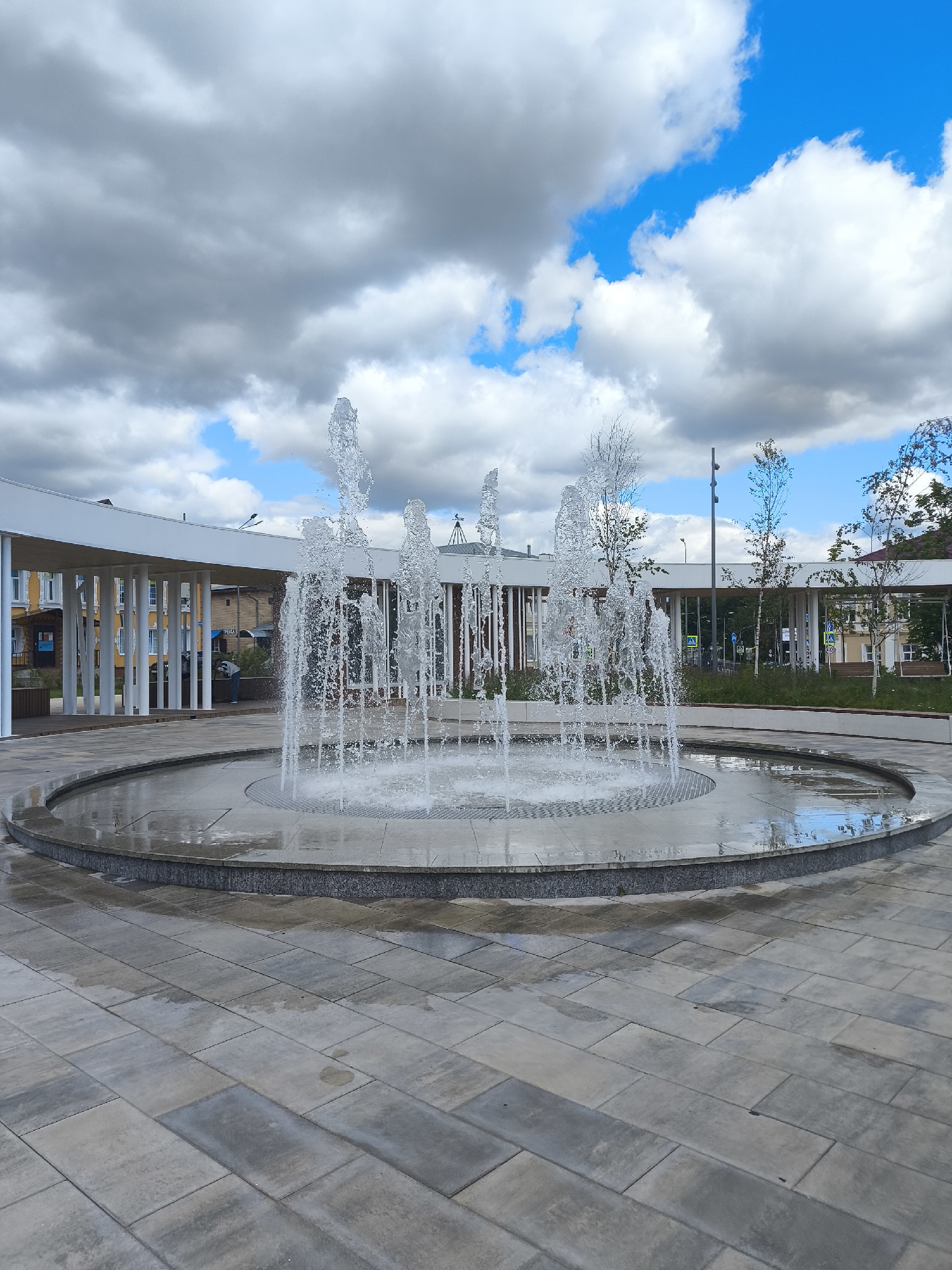 A medium shot captures a modern water fountain in an outdoor public space. The fountain, located in the foreground, features multiple jets of water arcing upwards and outwards from a circular basin. The basin itself is surrounded by a paved area with large, rectangular grey stones, some of which are wet, reflecting the sky. In the mid-ground, a curved, white, open-air structure with numerous white pillars provides shade. Behind this structure, a few trees with sparse foliage are visible. Further in the background, buildings with yellow and white facades and windows line a street. The sky above is a mix of grey clouds and patches of bright blue, with some white cumulus clouds. The overall scene appears to be a contemporary urban plaza, likely in Taldom, Russia, designed for public recreation. There is one person visible in the background, wearing a blue shirt and jeans, standing near the white structure.