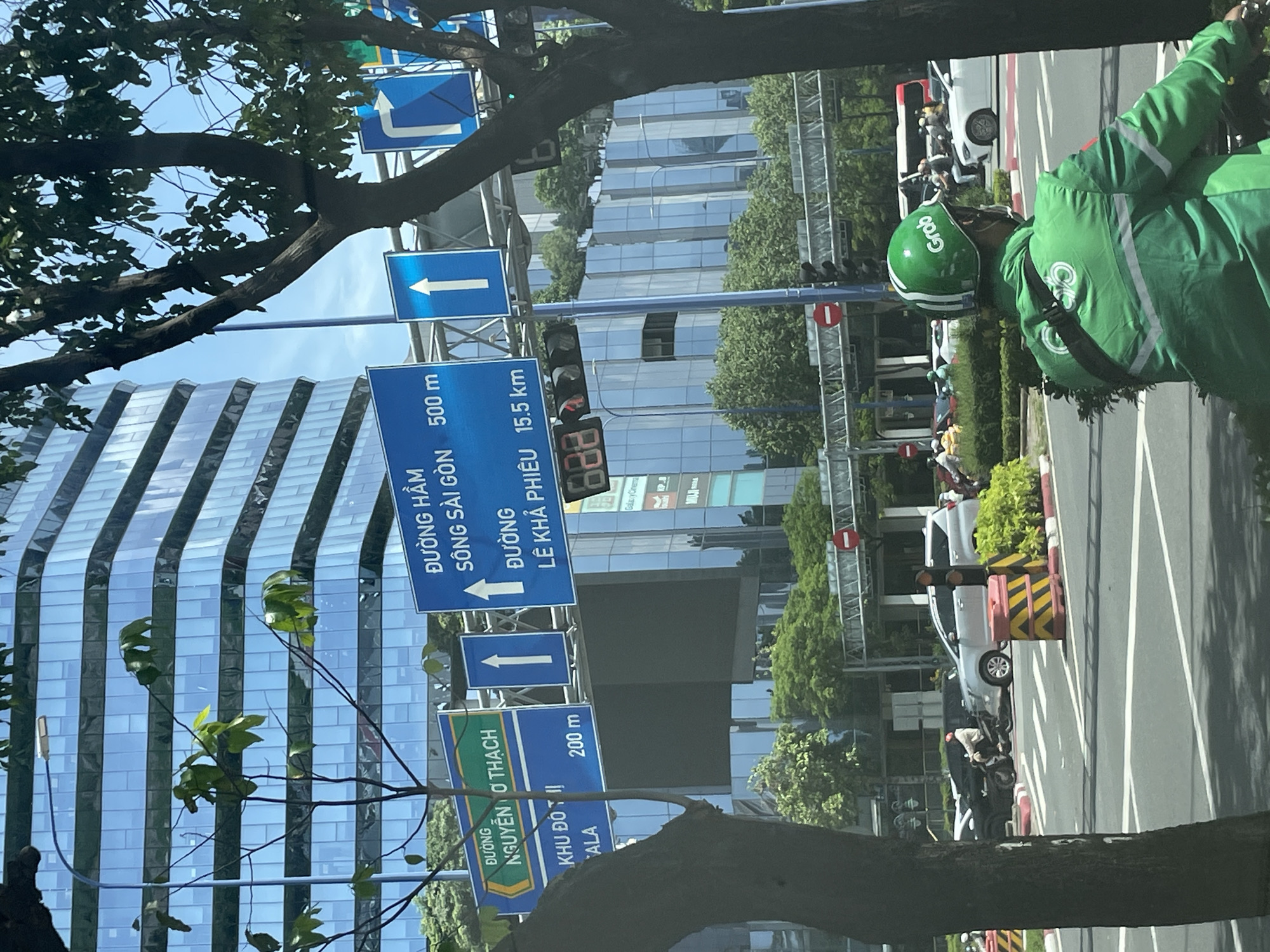 The image captures a street scene in Thủ Đức City, Vietnam. A Grab delivery rider, wearing a green helmet and jacket with the Grab logo, is in the foreground, facing away from the camera. The rider is on a motorcycle, suggesting they are actively working. Behind the rider, a busy intersection is visible with several vehicles, including cars and motorcycles, navigating the road.
Several blue directional signs are mounted on poles. One sign indicates "ĐƯỜNG HÀM SÔNG SÀI GÒN 500 m" and "ĐƯỜNG LÊ KHẢ PHIÊU 15.5 km". Another green and blue sign reads "ĐƯỜNG NGUYỄN O THẠCH" and "KHU ĐÔI TI 200 m". Traffic lights, including a digital display showing the time "888", are also present.
The background is dominated by a modern glass-fronted skyscraper with multiple levels of reflective windows. Lush green trees and shrubs line the roadside, providing a contrast to the urban architecture. The overall scene depicts a vibrant urban environment with active transportation and commercial activity.